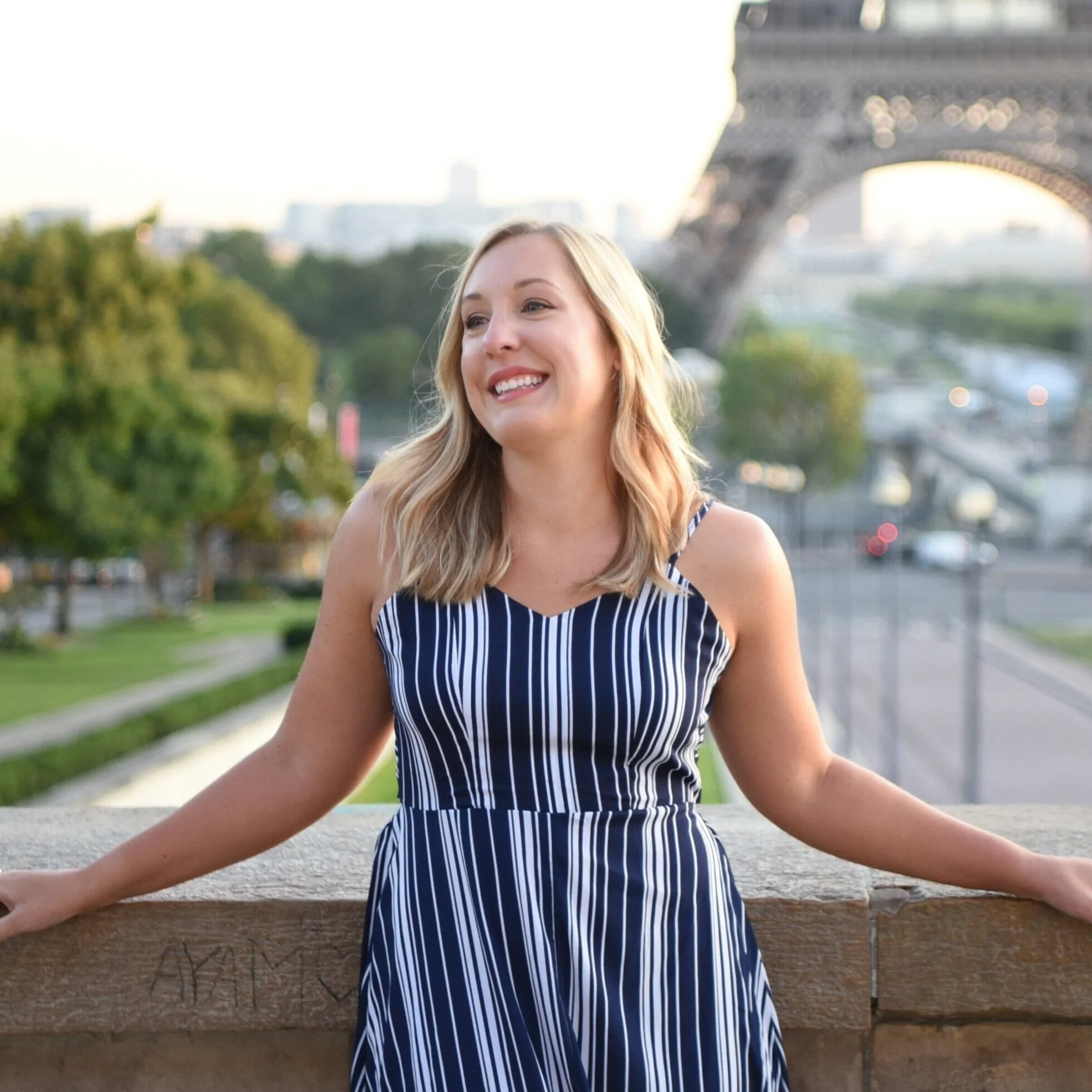 A young woman with long blonde hair smiling, sitting outdoors on a stone bench, with the Eiffel Tower in the background, during daylight hours.