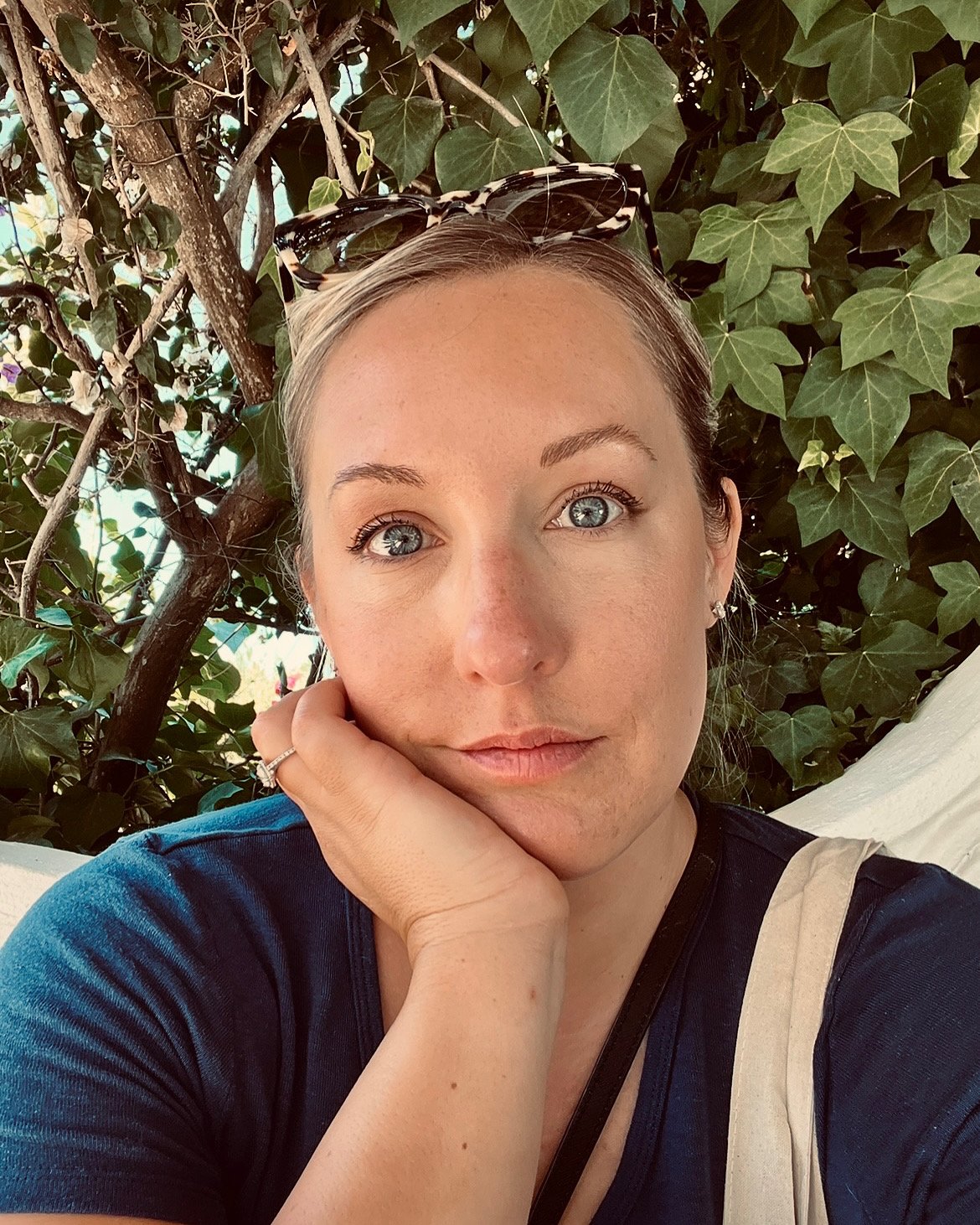 A woman with blonde hair and blue eyes resting her face on her hand, wearing sunglasses on her head, a navy blue shirt, and a beige strap over her shoulder, sitting in front of green ivy and tree branches.
