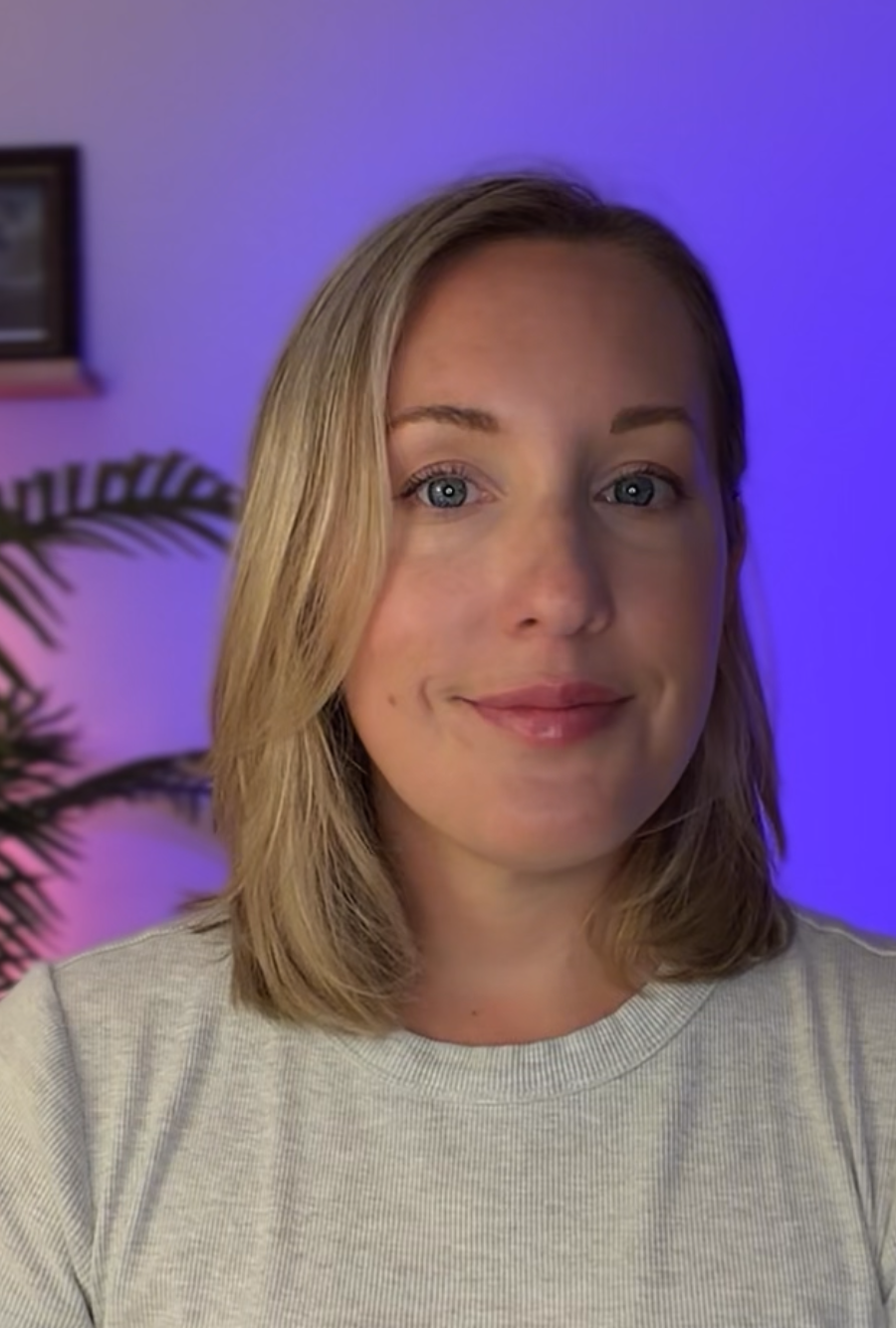 A woman with shoulder-length blonde hair and blue eyes smiling at the camera, wearing a light-colored shirt, with a purple-lit background and a plant to her left.