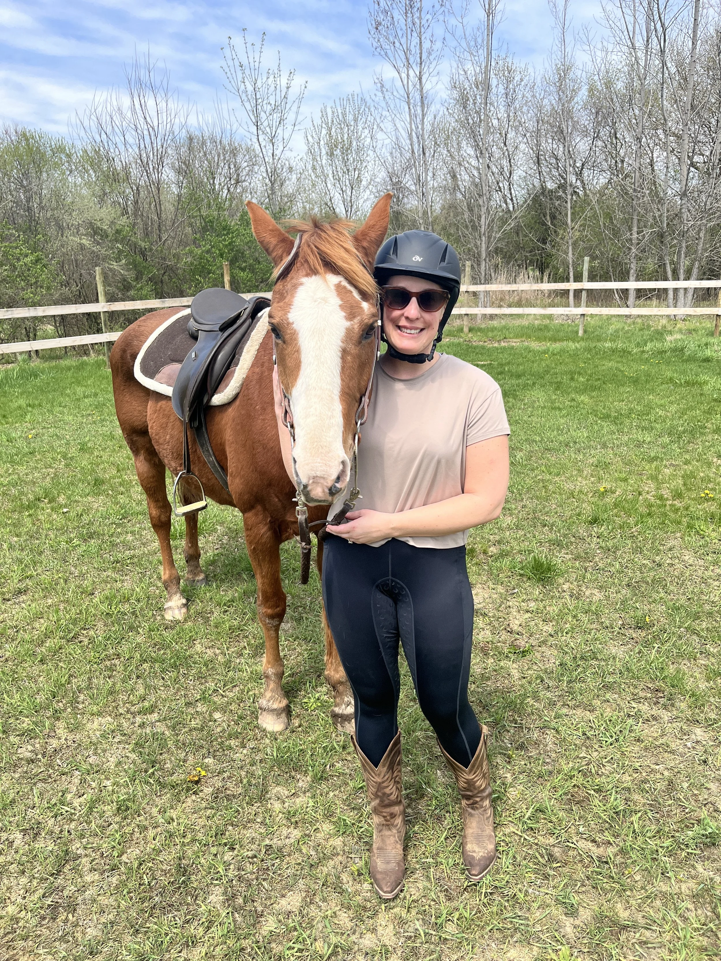 A woman wearing a black riding helmet, sunglasses, a beige t-shirt, and black riding pants, standing next to a brown horse with a white blaze on its face, in an outdoor grassy area with a wooden fence and trees in the background.