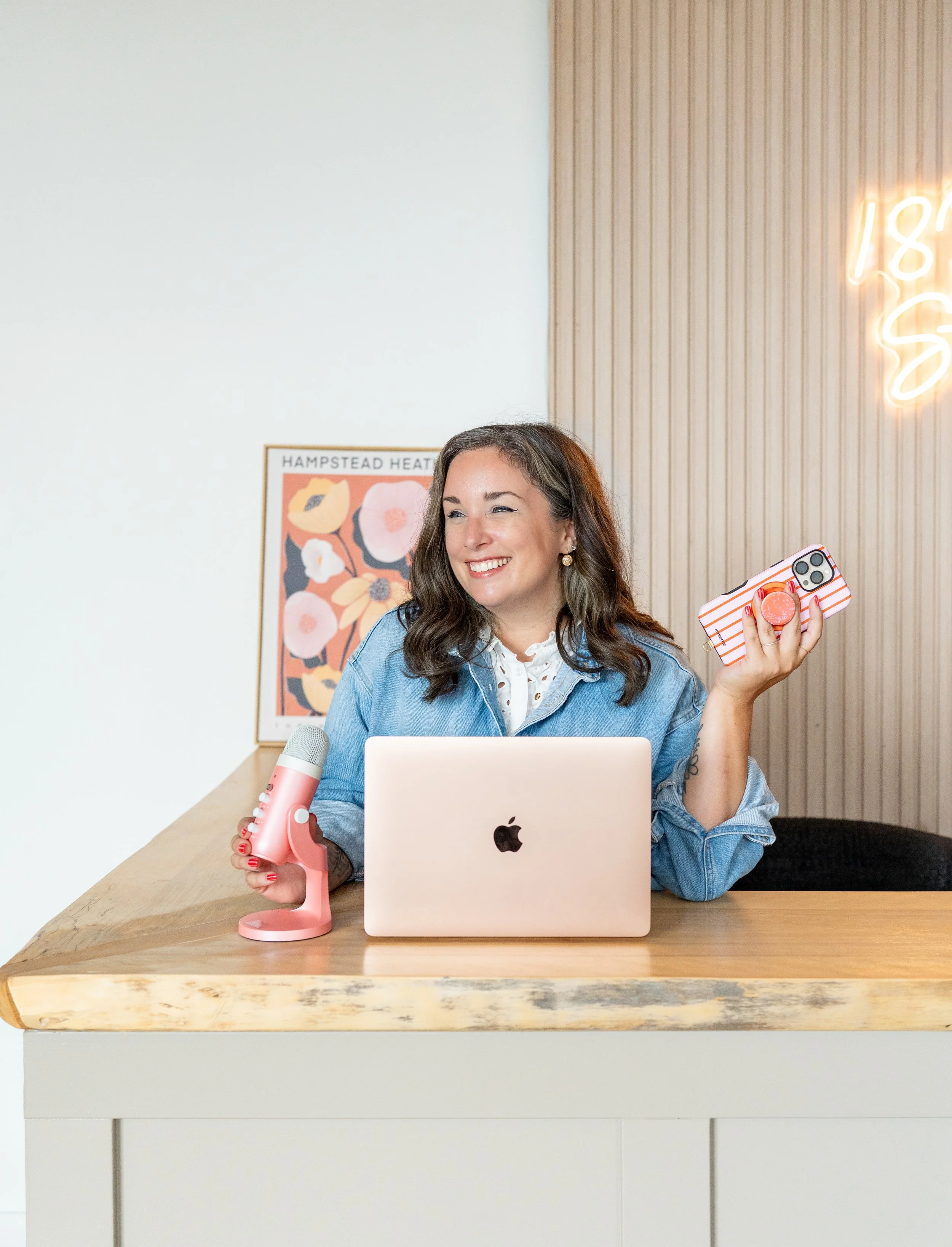 A smiling woman sitting at a wooden table in front of a silver laptop, holding a pink phone with a striped case and a pop socket, with a pink microphone on the table, in a modern room with a poster that says 'Hampstead Heath' and a neon sign on the wall.