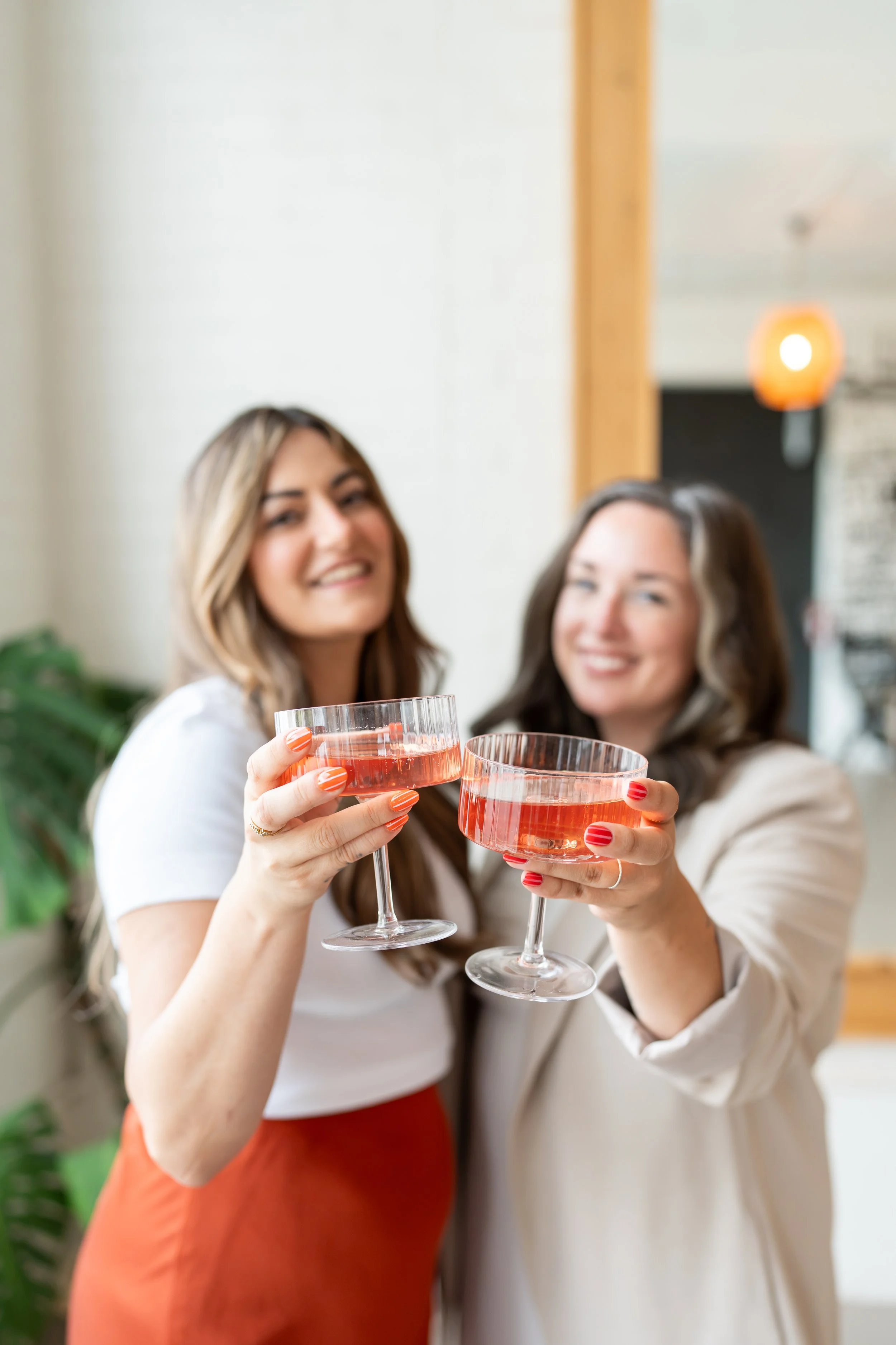 Two women smiling and clinking glasses of pink wine in a casual indoor setting.