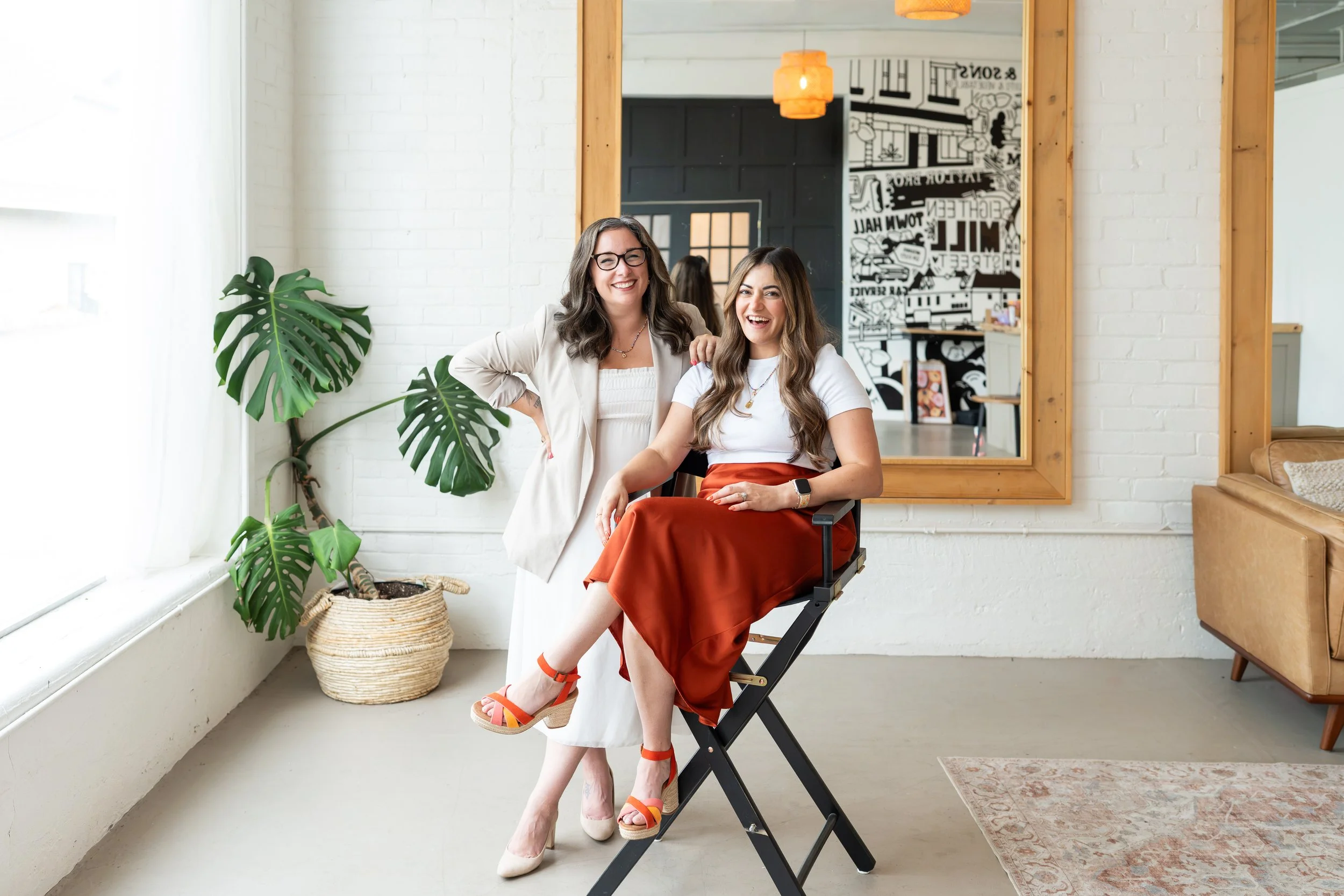 Two women smiling and posing inside a bright room with white brick walls and large window. One woman seated in a director's chair, wearing white top, rust-colored skirt, and orange sandals. The other woman standing beside her, wearing glasses and a beige jacket. Green plant and wall art visible in the background.