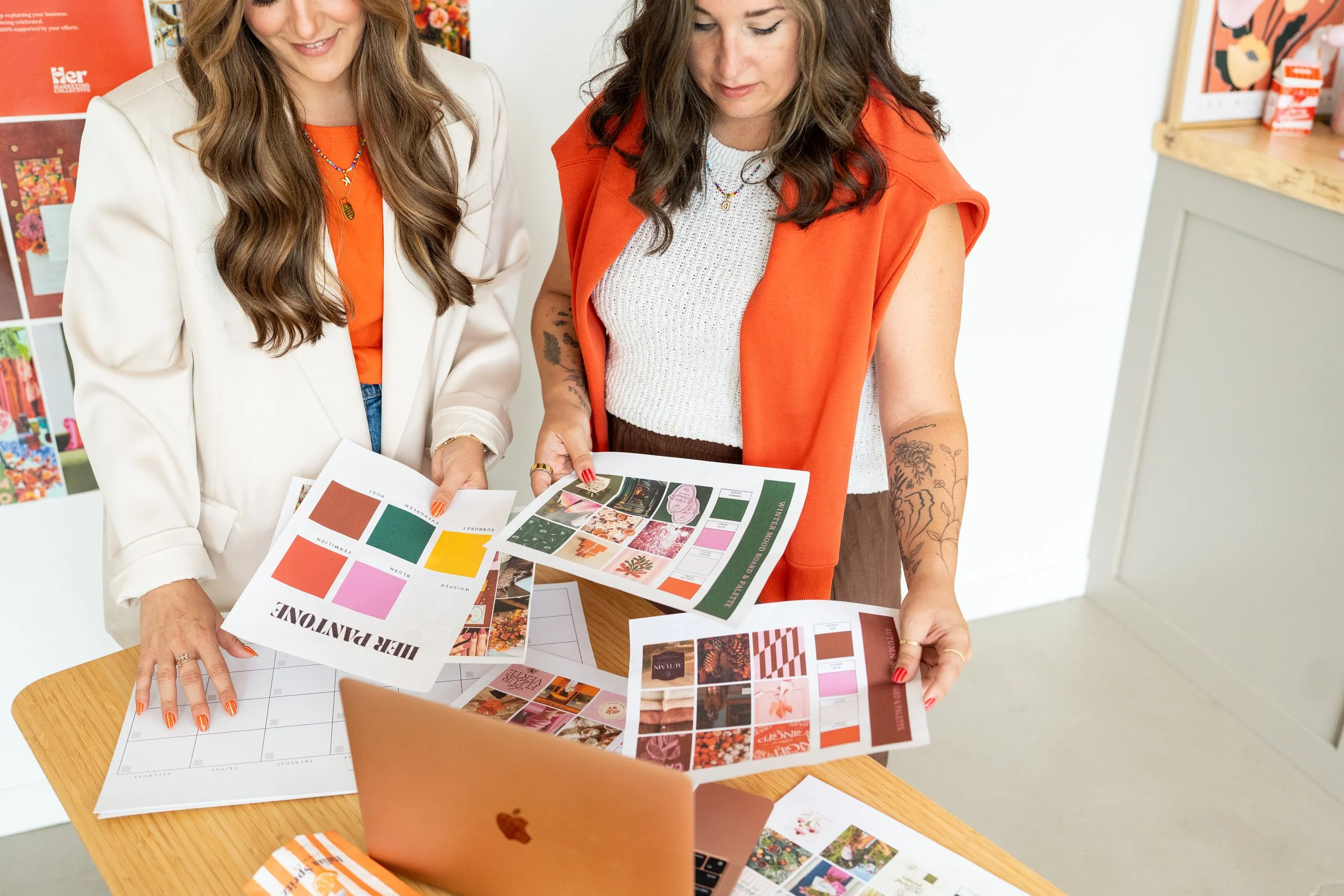 Two women are looking at printed design sheets on a wooden table, with a laptop and colorful arts and crafts materials nearby in a bright workspace.
