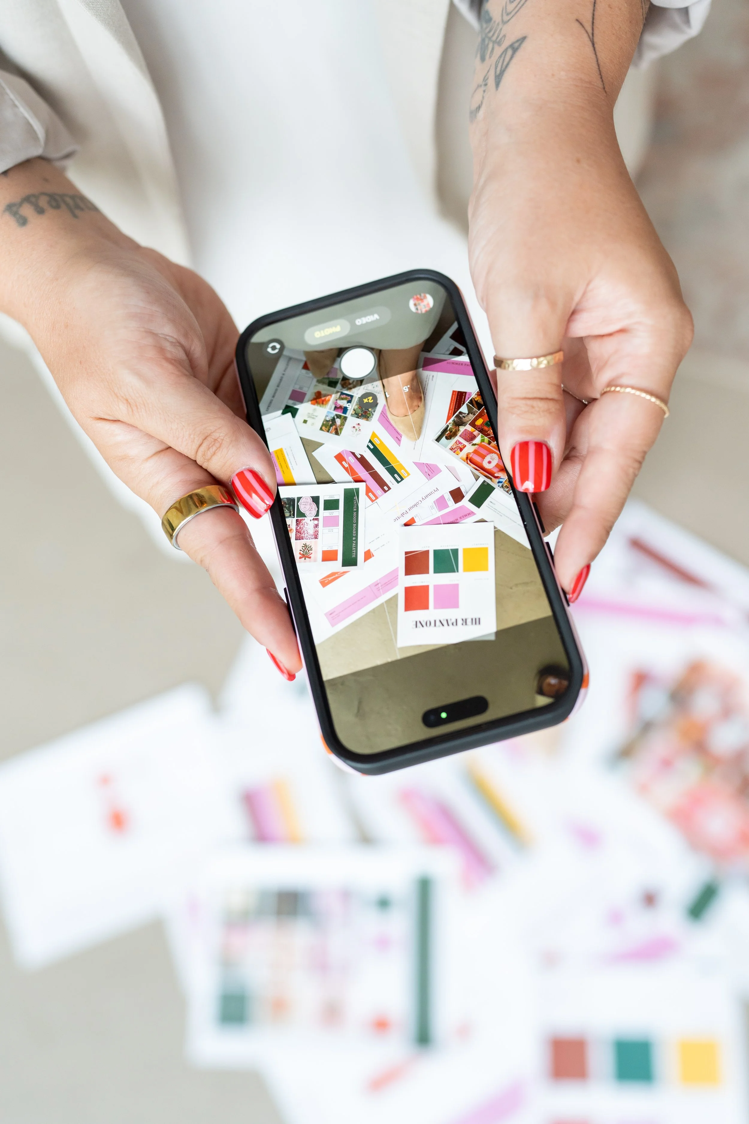 A person with painted nails holding a smartphone, taking a picture of a flat lay display. The display contains color swatches and design samples on sheets of paper.