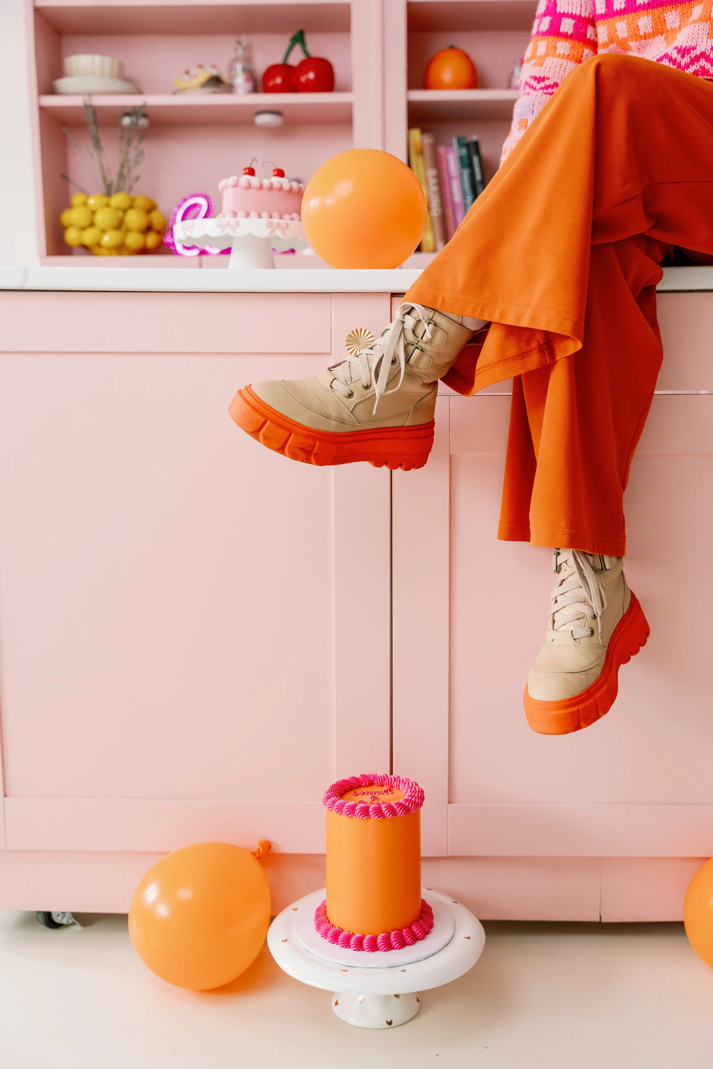 A person sitting on a pink kitchen counter with legs dangling, wearing beige and orange platform shoes and orange wide-leg pants, with a pink birthday cake, orange balloons, and colorful food items in the background and foreground.