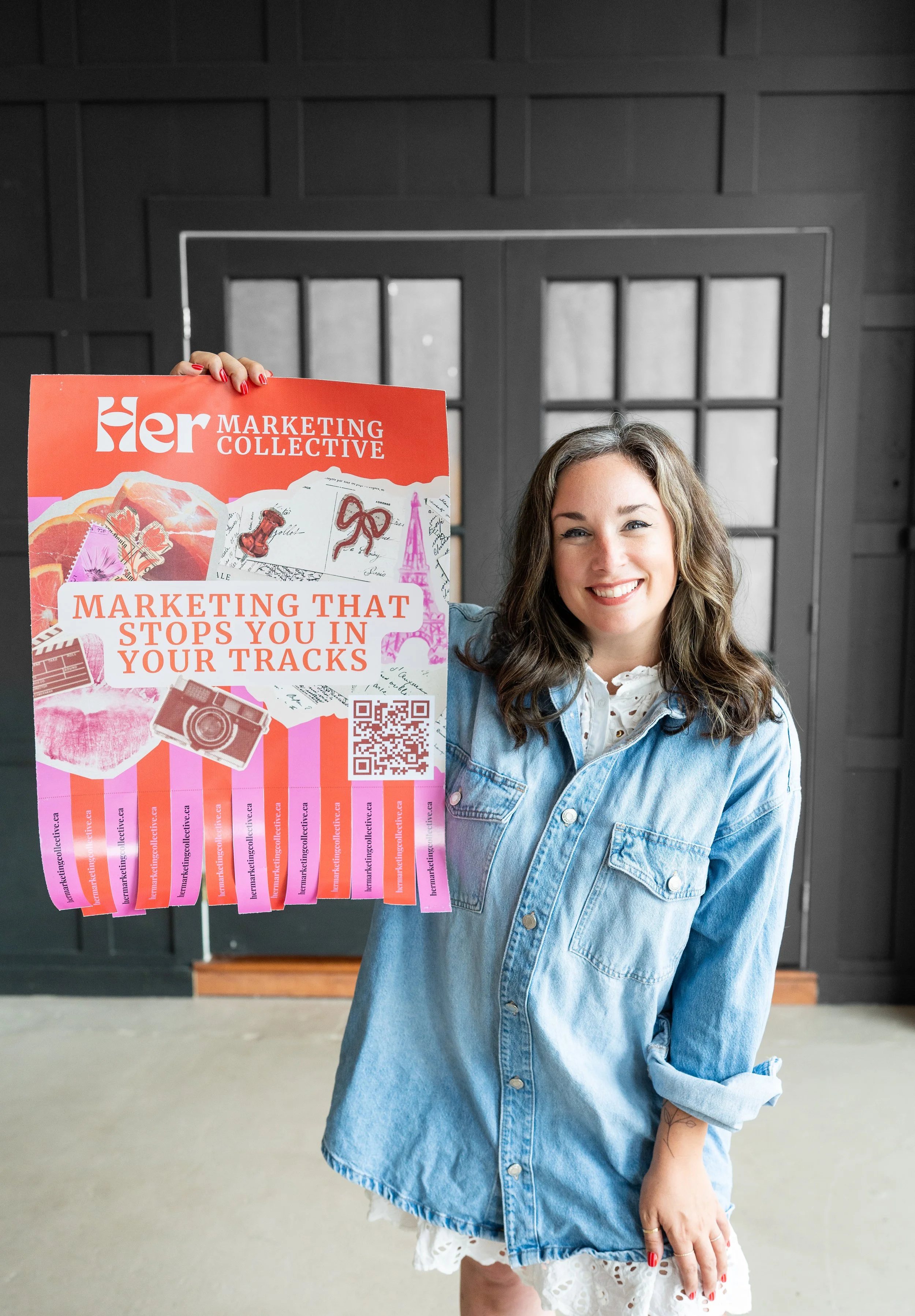 A woman with wavy brown hair, light skin, and red painted nails smiling and holding a promotional poster for Her Marketing Collective in front of a black wall and glass double door.