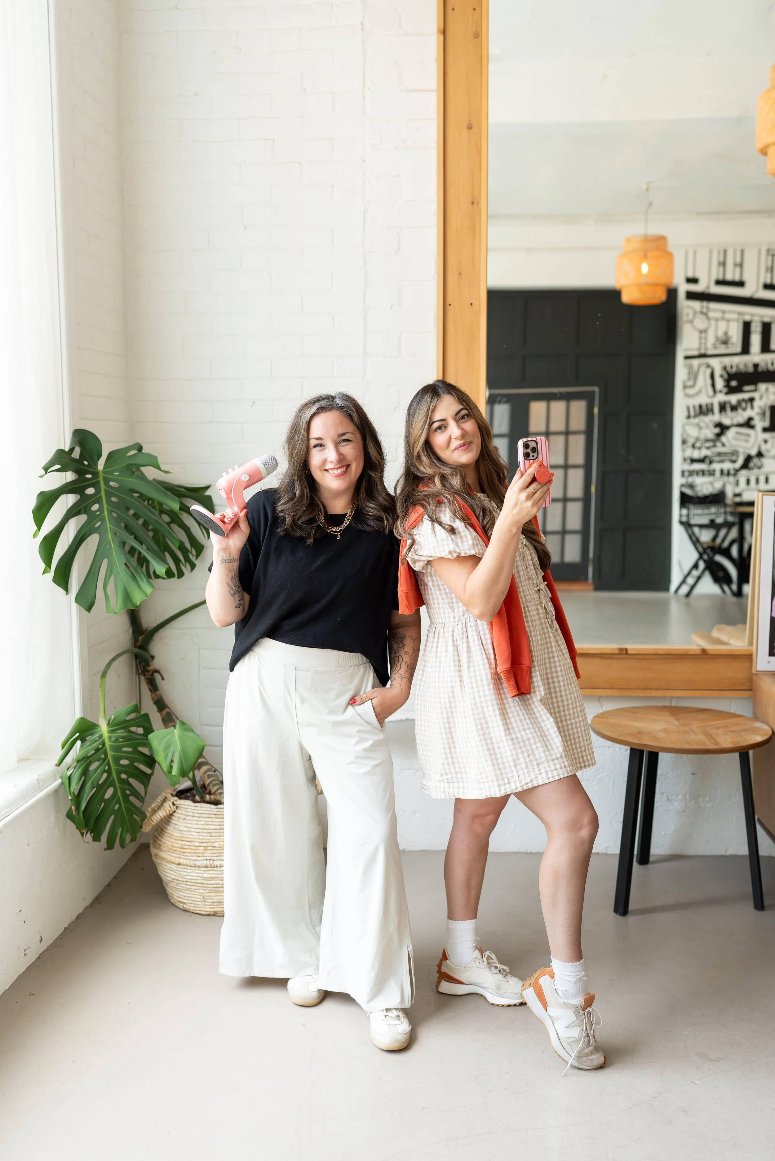 Two women posing indoors, one holding a pink hairdryer and the other taking a selfie with a smartphone, in a room with white brick walls and a large green plant.