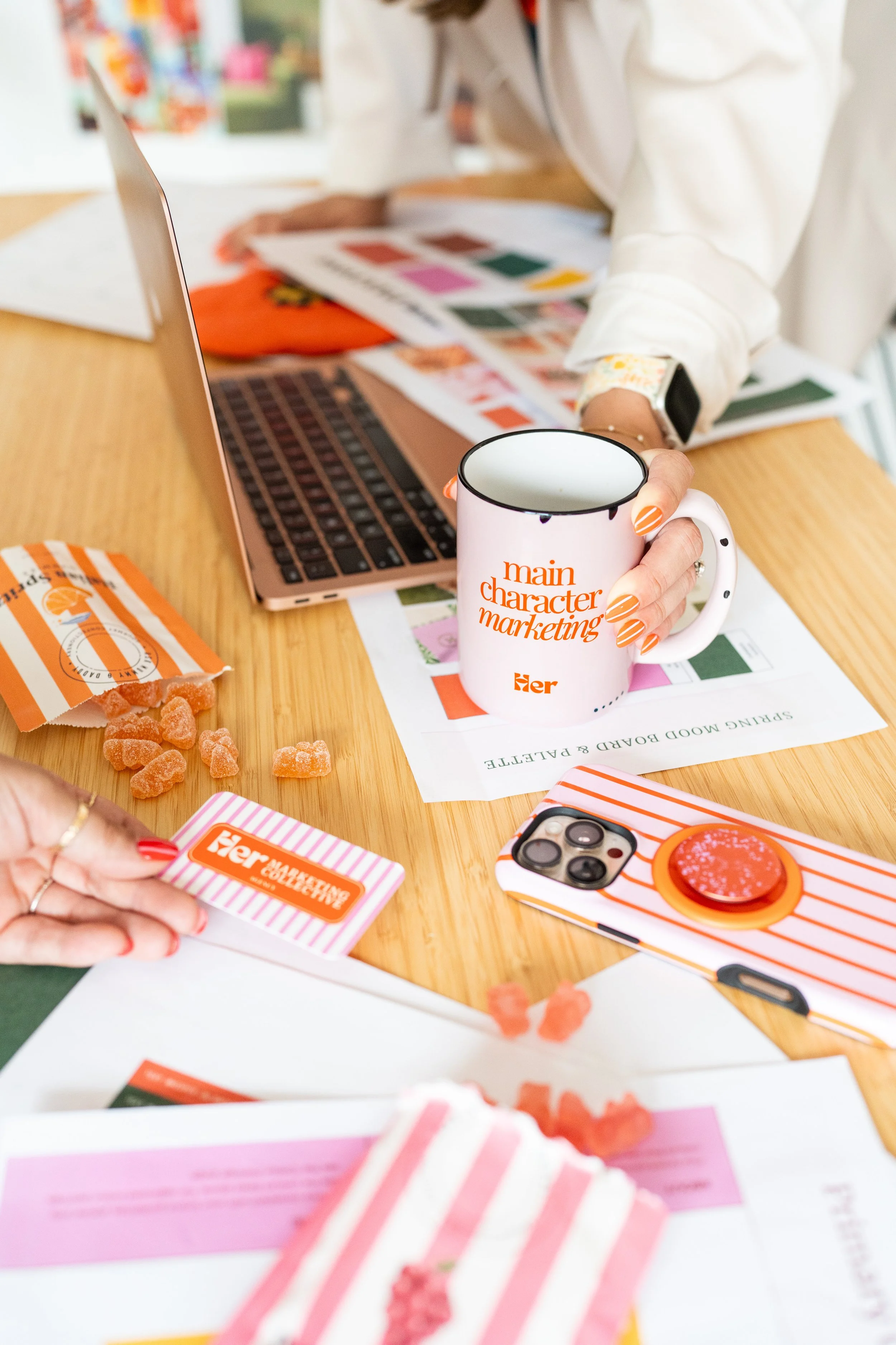 Close-up of a person holding a pink and orange card with the text 'her marketing' on it, with a coffee mug and phone on a table, and other paper materials and orange gummy candies around.