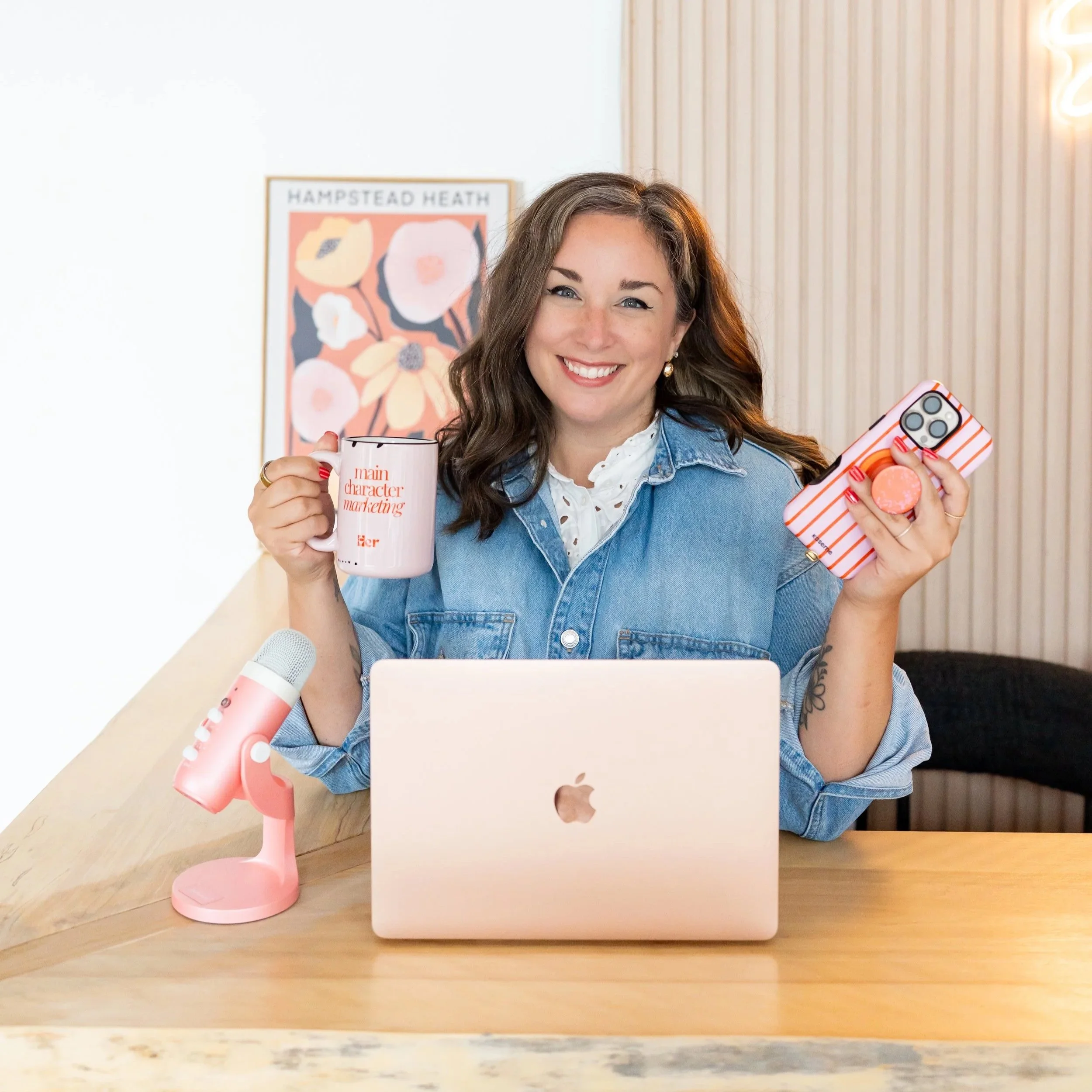 A woman with brown hair and a denim jacket smiling while holding a pink mug and a striped phone case, sitting at a wooden table with a white tablet, in a cozy room with a wall and a poster in the background.