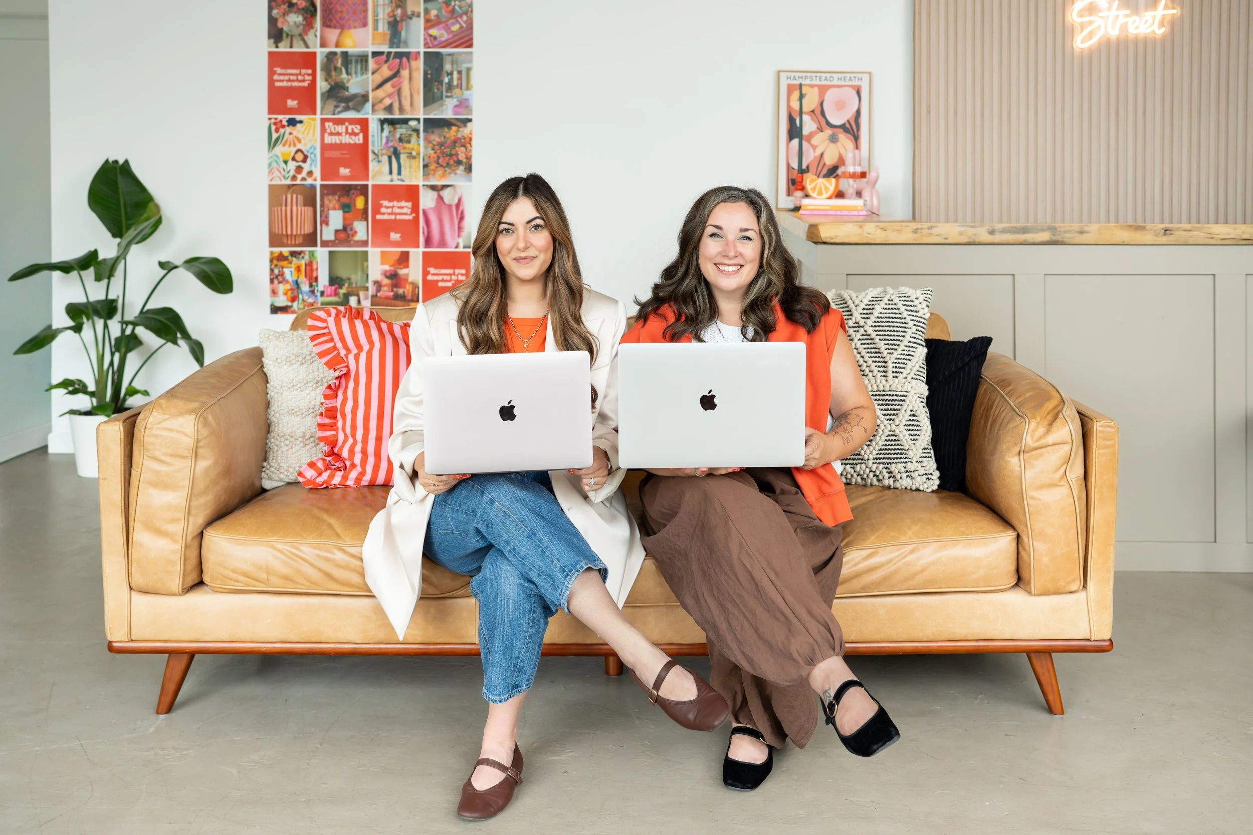 Two women sit on a beige leather sofa in a modern, stylish office or creative workspace, each working on a silver Apple MacBook. They are smiling and dressed casually; one with long brown hair in a white blazer, and the other with curly dark hair in an orange top and brown pants. The background features a collage of colorful images on the wall, a large green houseplant, and decorative items including a neon sign that says 'Street' and framed art.