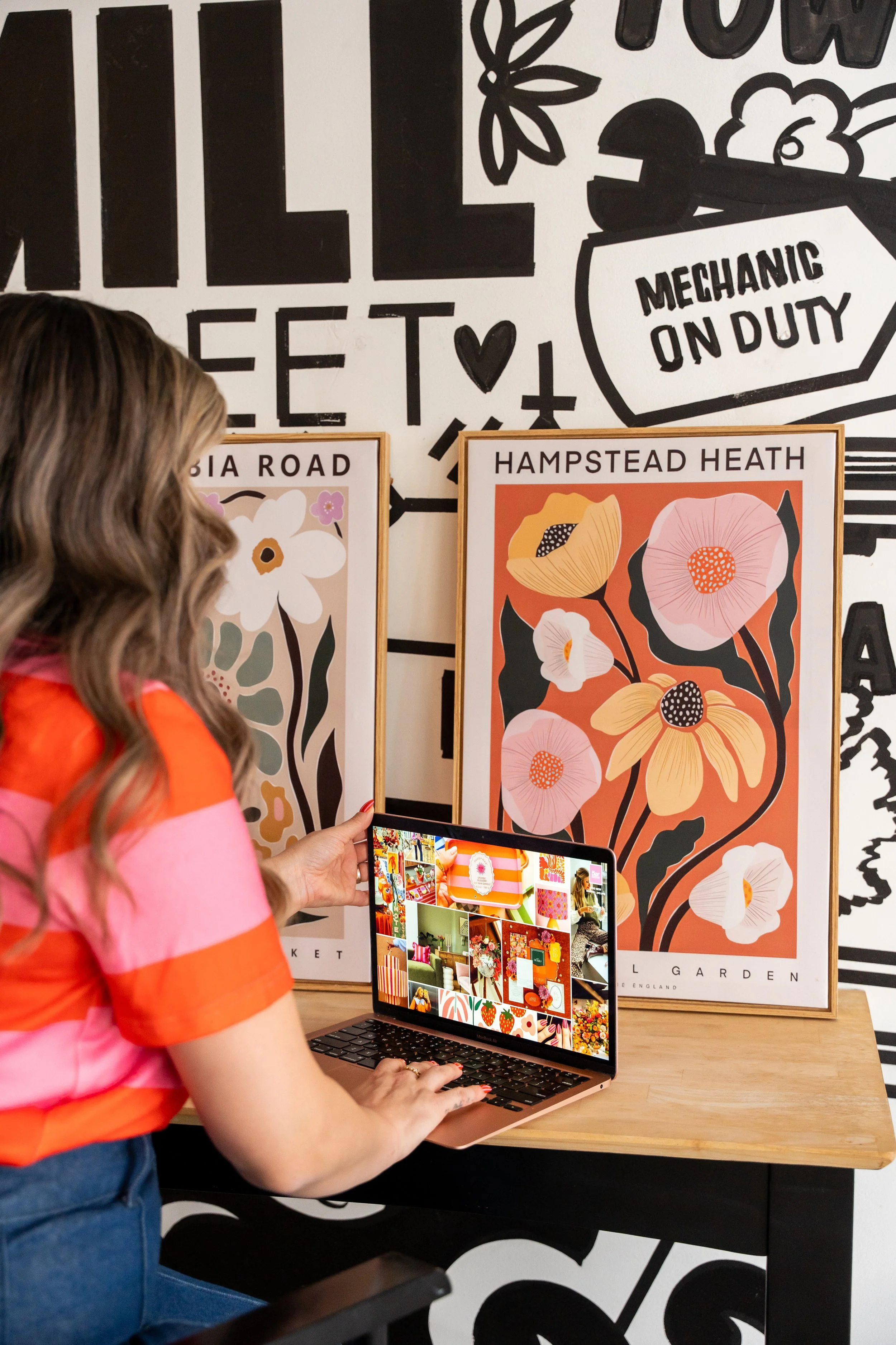 A woman is taking a photo of a colorful floral poster with her phone. She is sitting at a wooden table, wearing a pink and orange striped shirt, and has wavy hair. Behind her, the wall has black and white decorative text and drawings related to a mechanical duty.