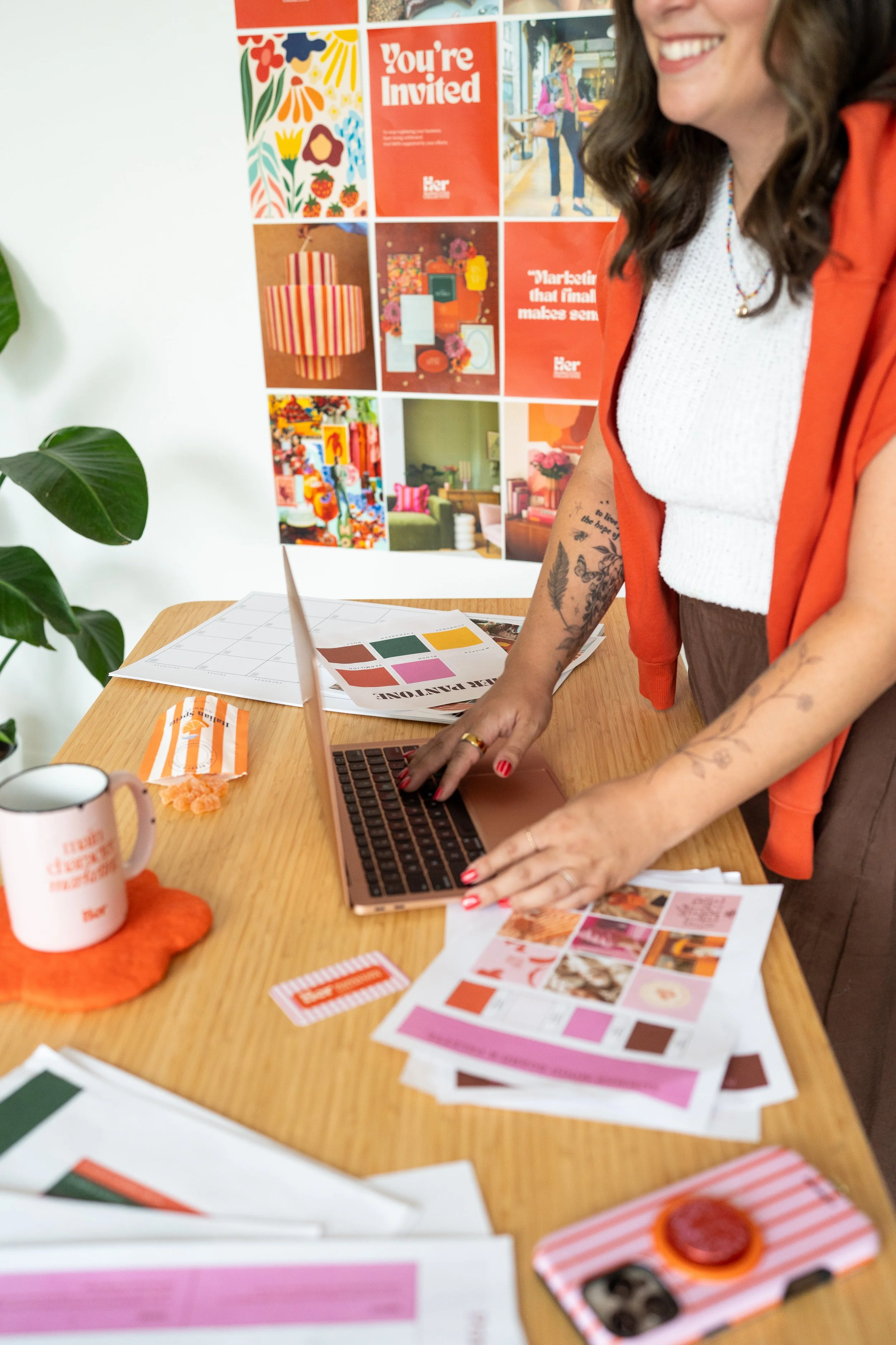 A woman working at a wooden table with colorful printed materials, a coffee mug, and a striped snack container, beside a green plant and a colorful collage on the wall.