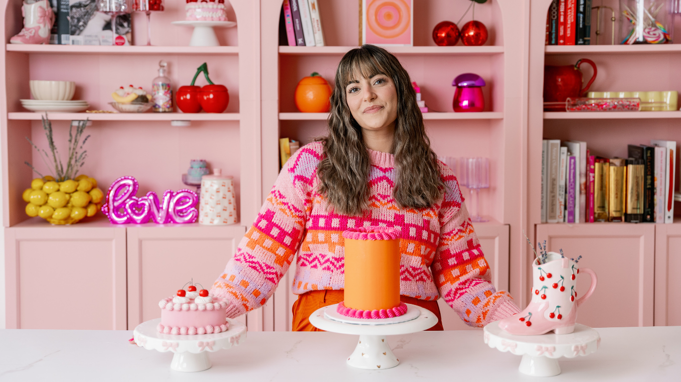 Woman in colorful sweater standing behind a table with three decorated cakes, pink background with shelves of books and decor, pink 'love' neon sign.