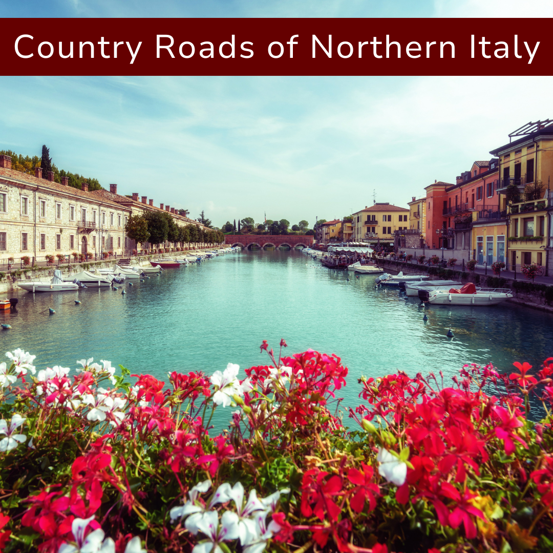 A scenic view of a canal in Northern Italy with colorful buildings on the right, boats docked along the edges, and a stone bridge in the background, framed by pink and white flowers in the foreground.