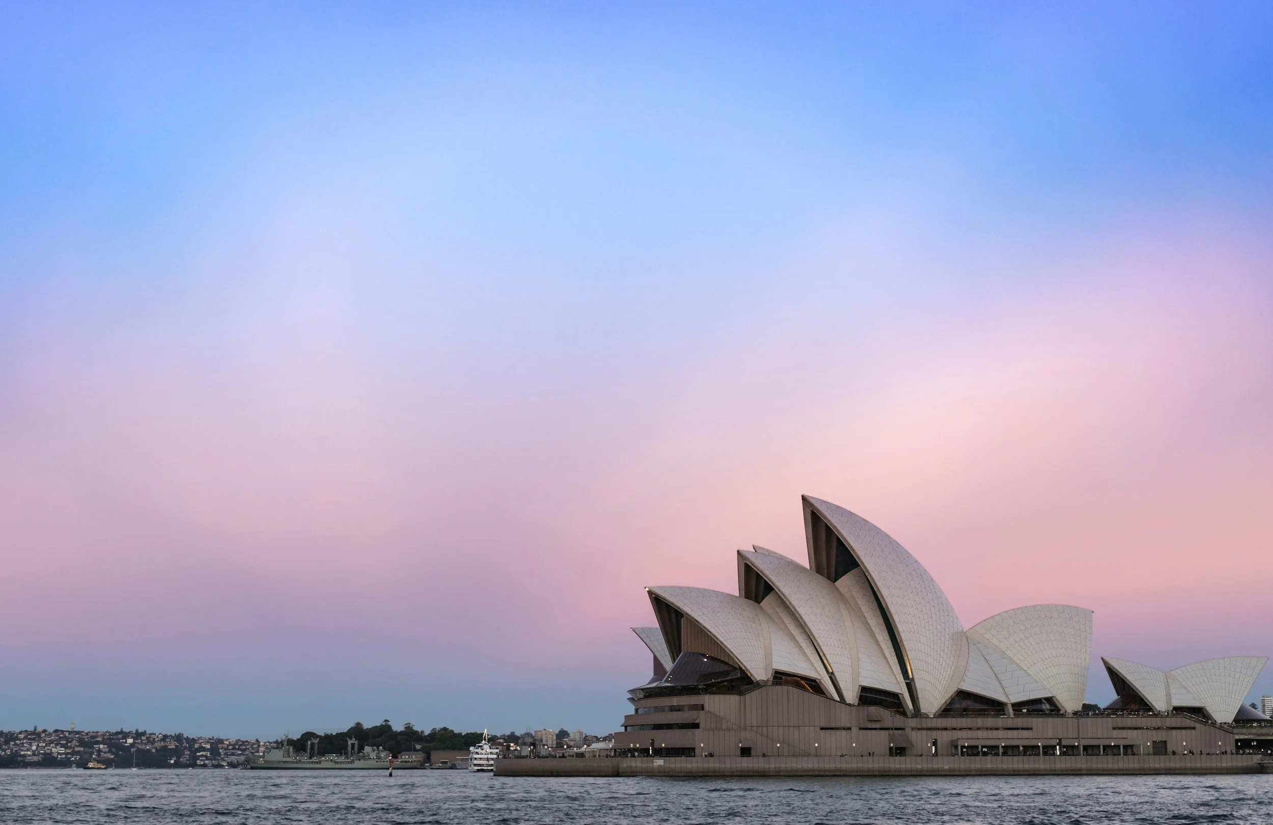 Sydney Opera House with a pastel pink and blue sky, viewed from across the water with boats in the harbor.
