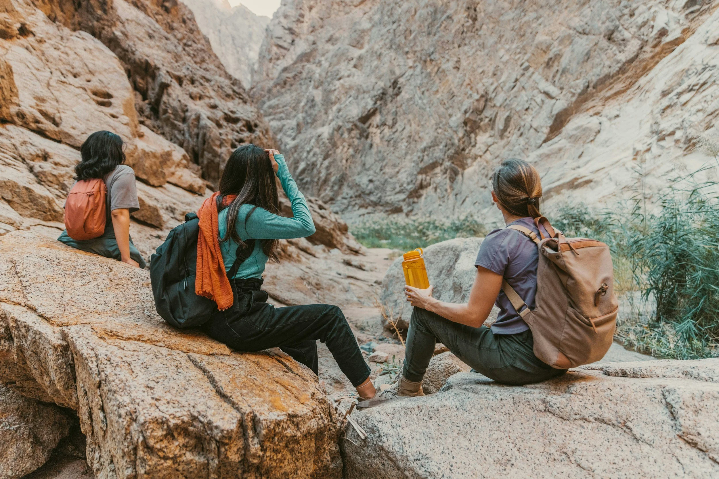 Three women with backpacks sitting on rocks and resting in a canyon surrounded by high rocky walls, one holding a water bottle.