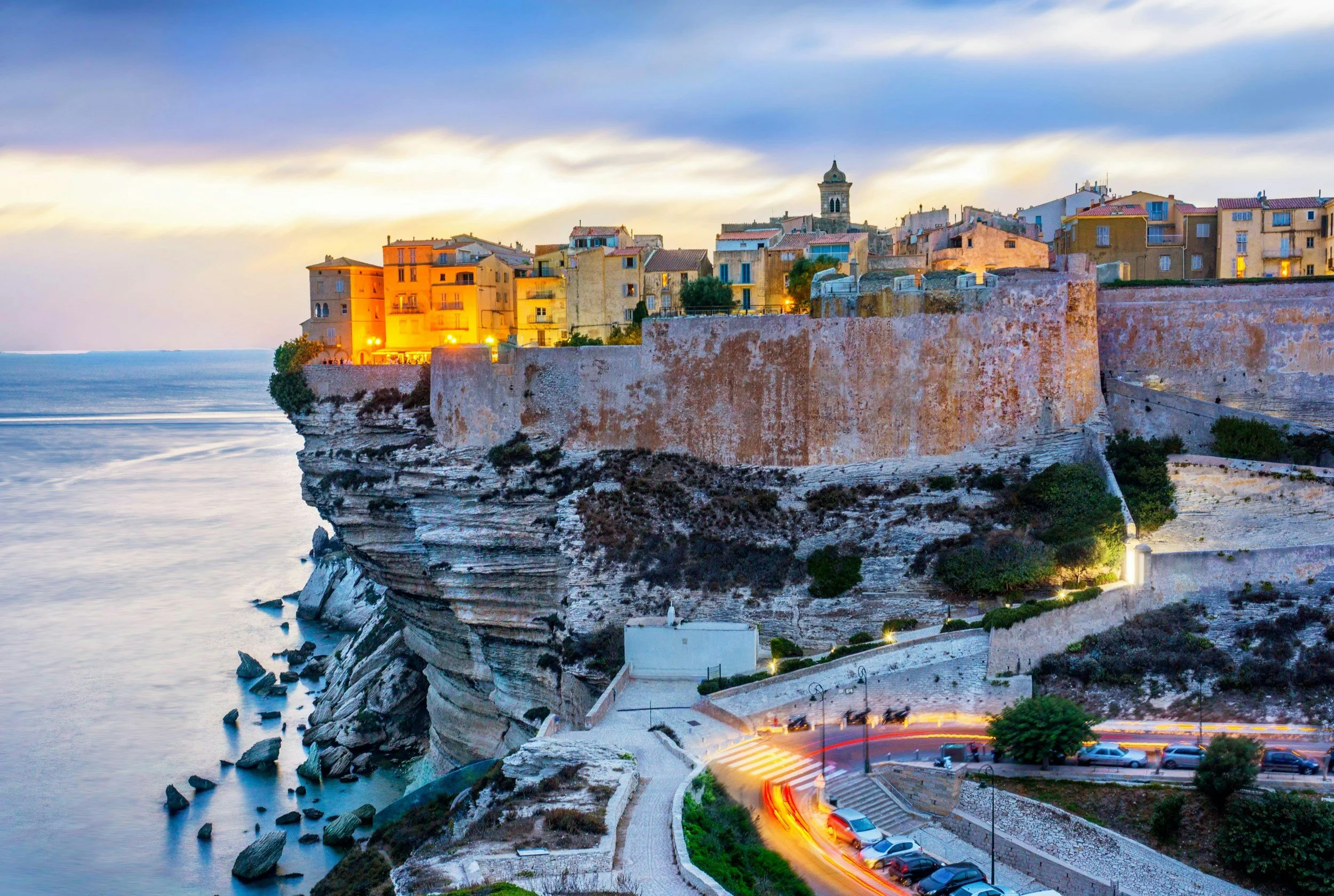 A coastal town at sunset with buildings on a cliff, a winding road with car light trails, and the ocean below.