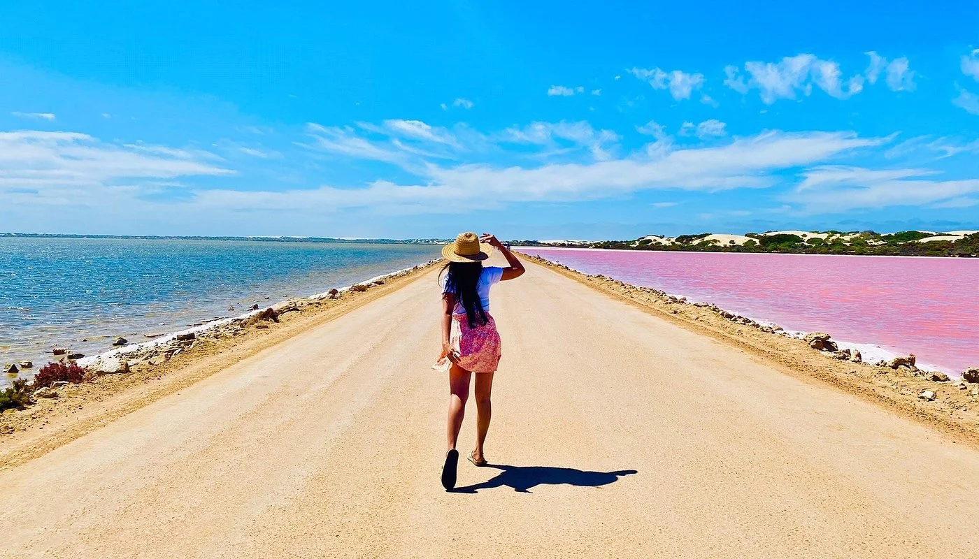 A woman walking along a dirt road between a blue water body on the left and a pink water body on the right under a blue sky with clouds, wearing a large straw hat, white top, and pink skirt.