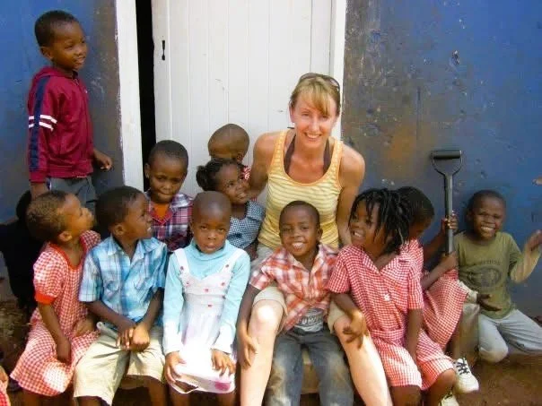 A woman surrounded by young African children, some in school uniforms, sitting and standing outdoors in front of a door and wall, smiling and enjoying the moment.