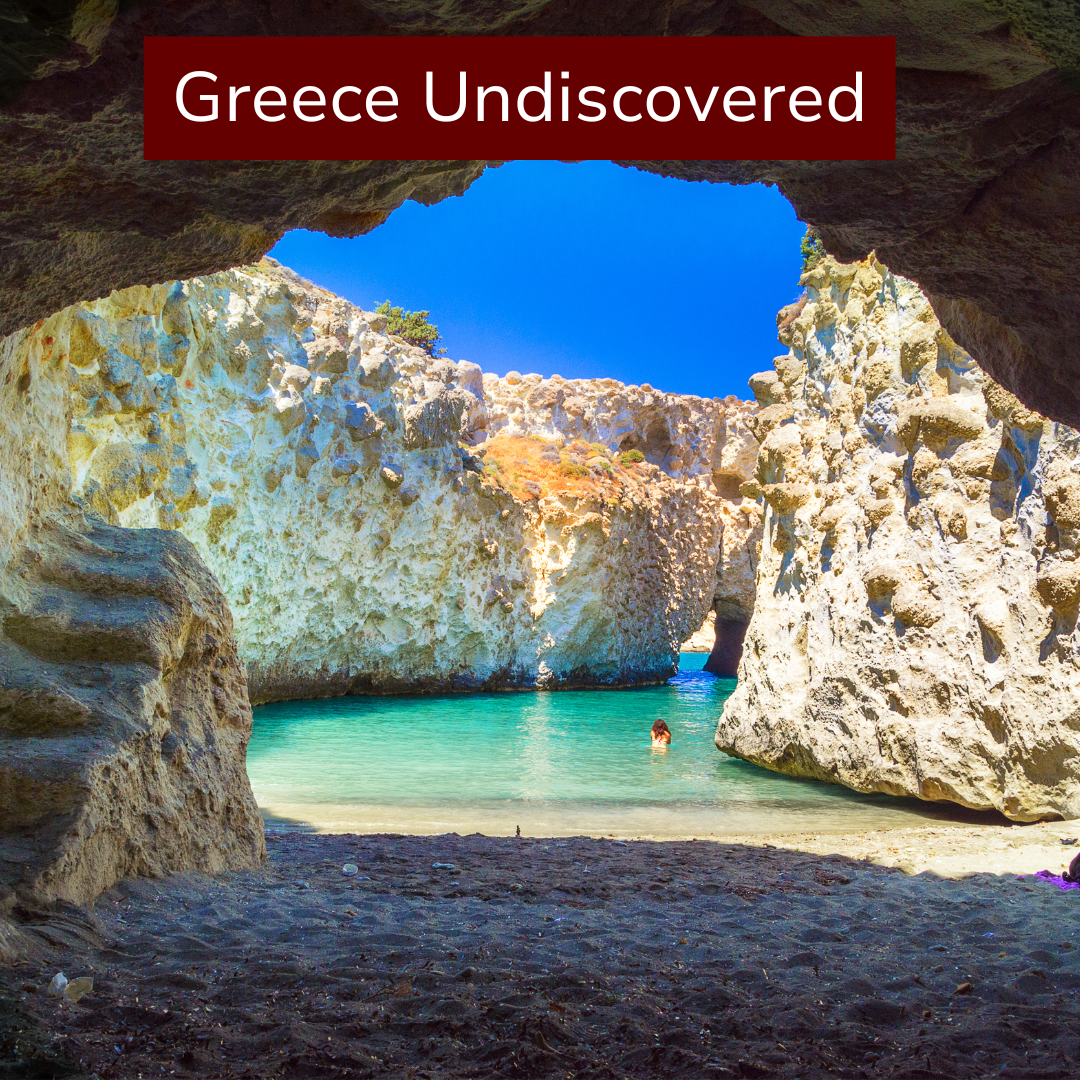 Sunlit cave opening revealing turquoise water and rocky cliffs on Greece's coast, with a person swimming and clear blue sky above.