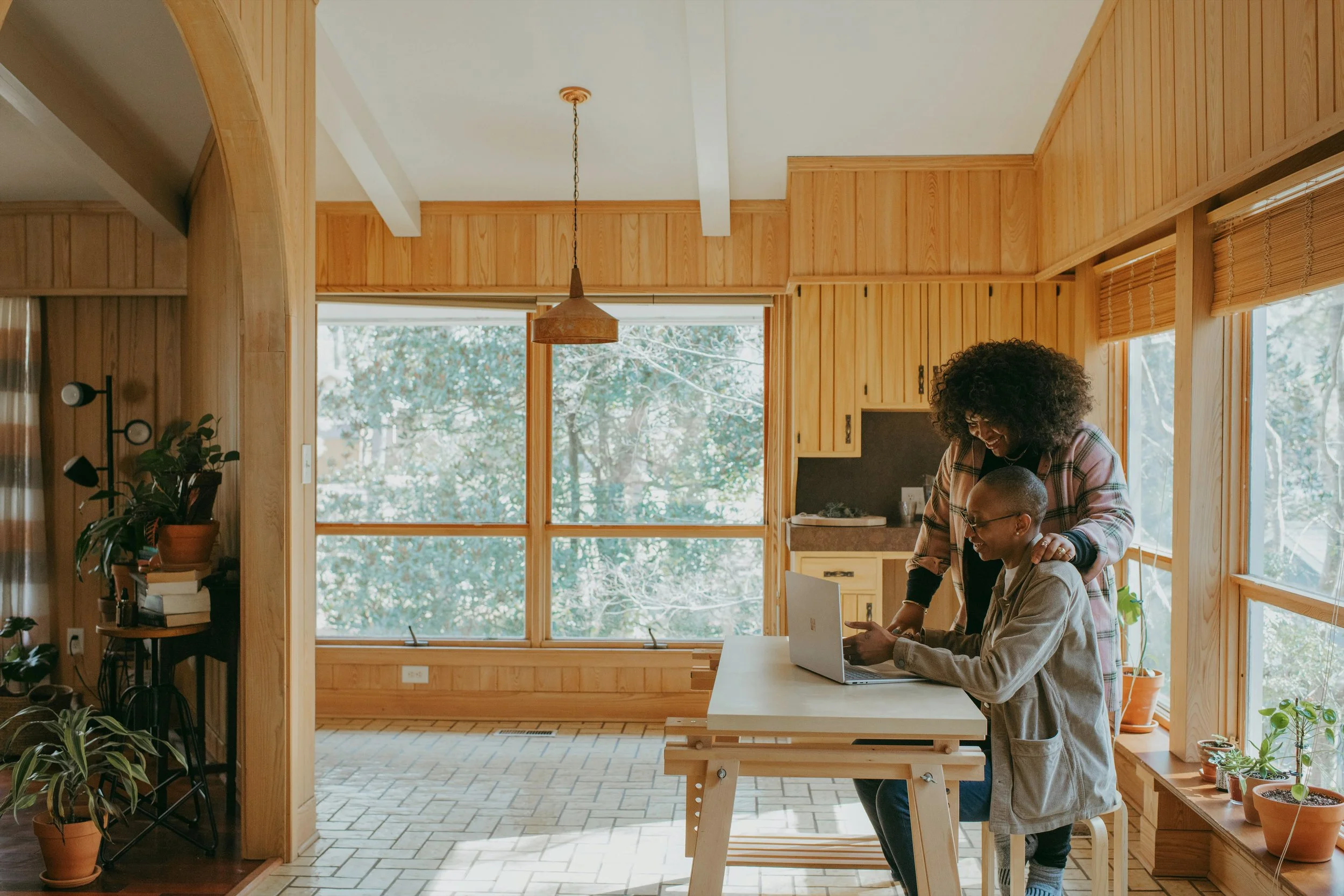 A woman and a person with glasses working on a laptop together in a wooden kitchen or dining area with large windows and potted plants.
