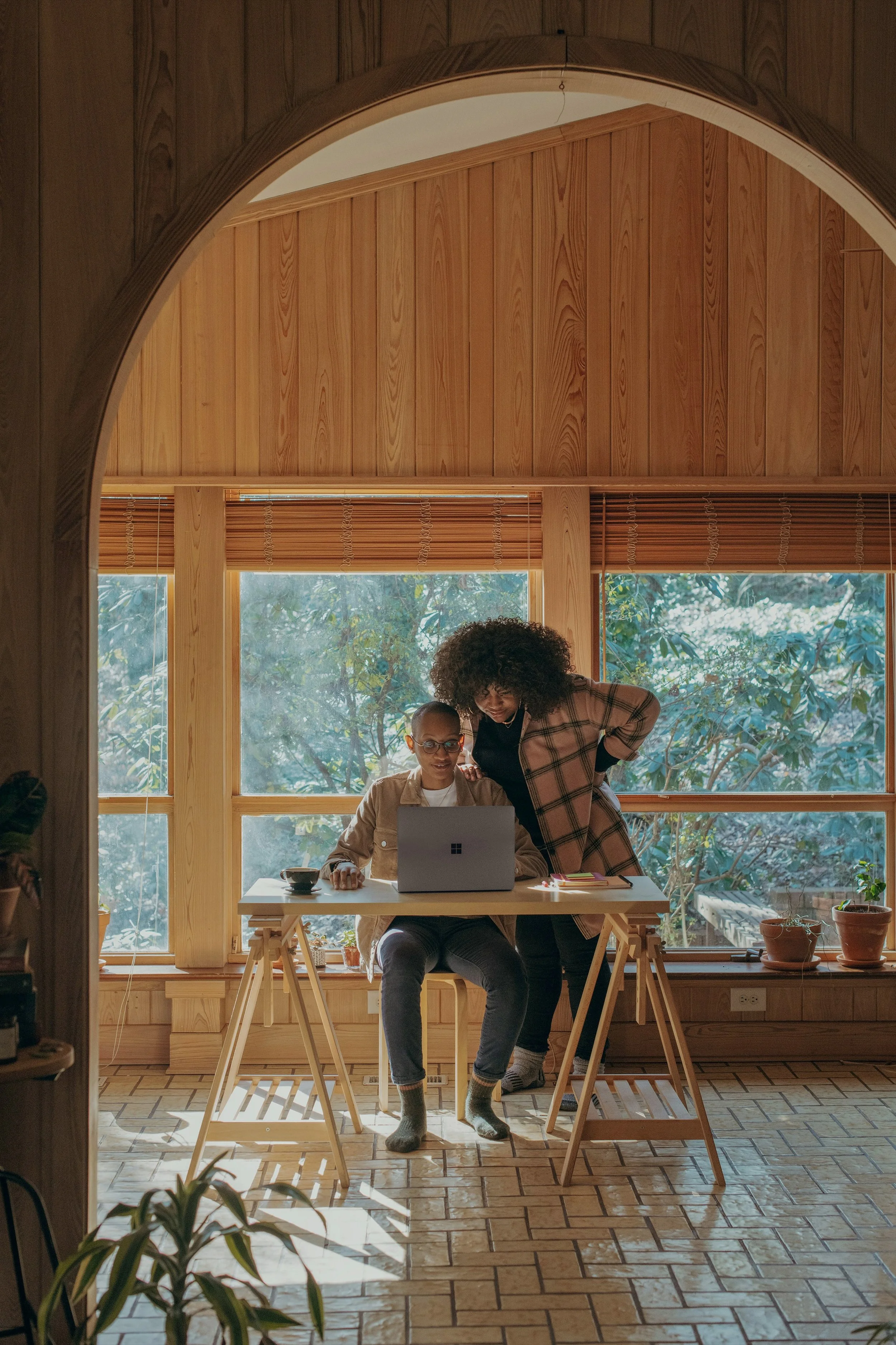Two women working together at a desk in a bright room with large windows and wooden walls.