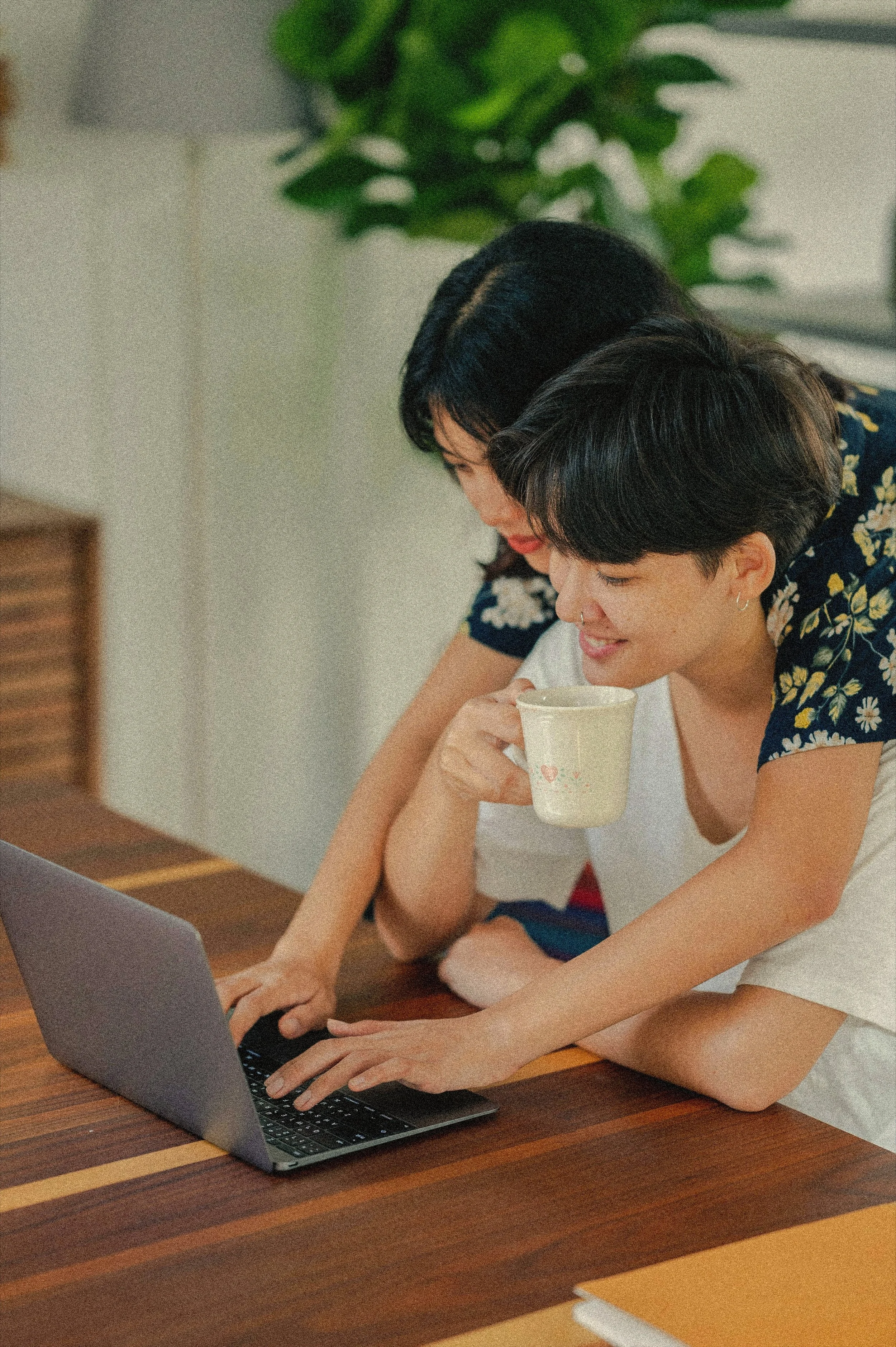 Two women are sitting at a wooden table, looking at a laptop screen together, with one woman holding a mug and smiling.