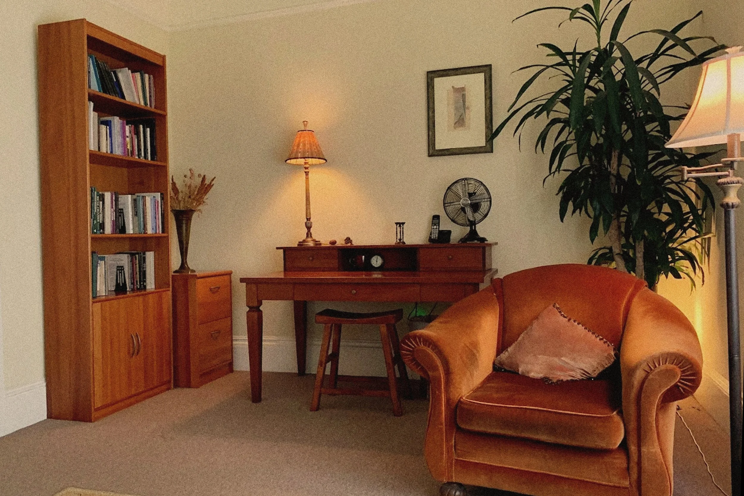 Living room with a wooden bookshelf filled with books on the left, a small wooden side table with dried flowers, a desk with a lamp, a vintage fan, and a clock, an orange velvet armchair with a pillow, a big potted plant, and a floor lamp on the right, beige walls, and framed artwork.