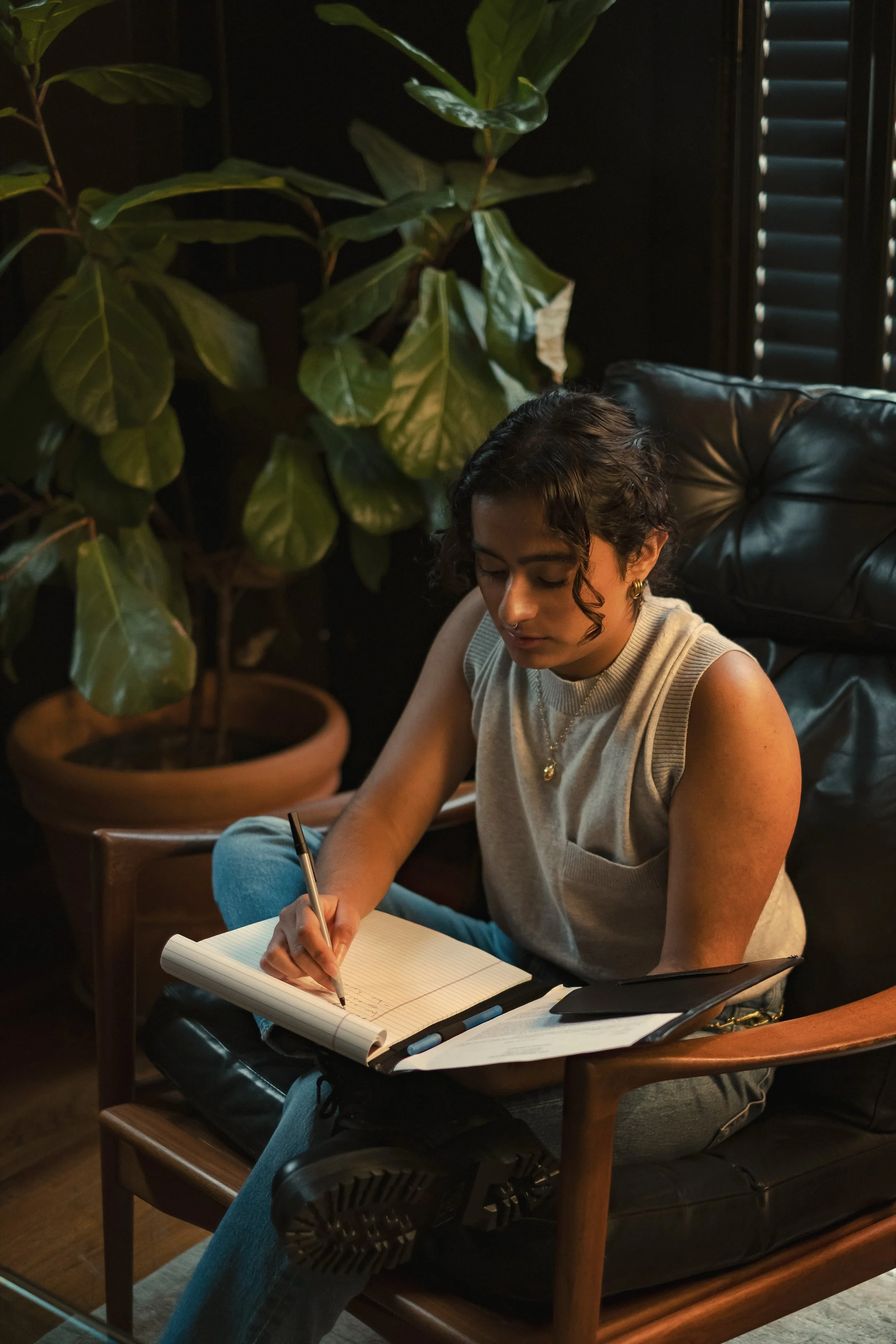 A woman sitting on a black leather armchair, writing in a spiral notebook with a black pen. She has short curly hair, multiple earrings, and is wearing a sleeveless beige top and blue jeans. There is a large potted plant behind her, and a window with black blinds to her right.