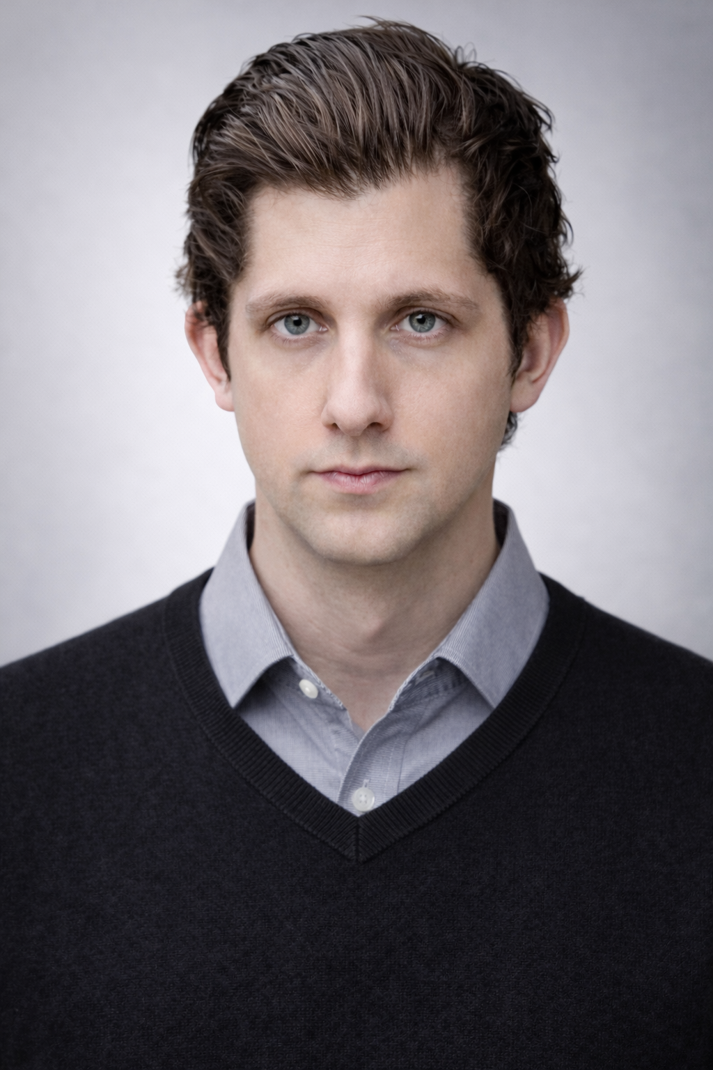 Headshot of a young man with brown hair, blue eyes, wearing a light gray collared shirt and a black sweater, posing against a plain gray background.