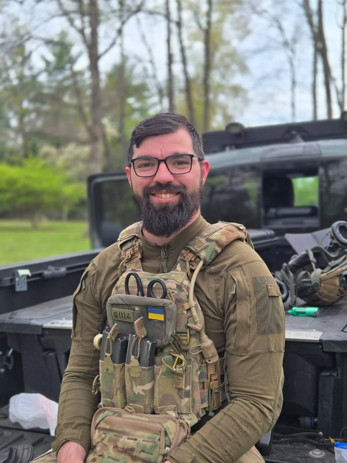 A smiling man in military camouflage gear standing outdoors near a black tactical vehicle with equipment on it, in a wooded area.
