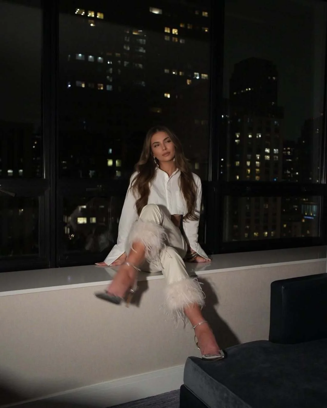 Woman in feather-trim cream pants and silver heels sitting on a high-rise window ledge overlooking Chicago at night.