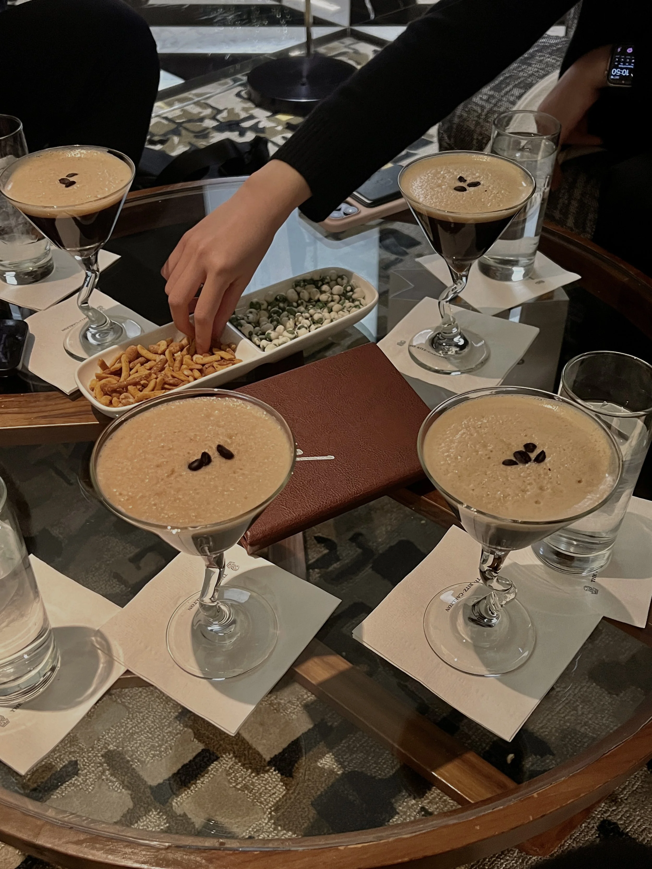 Four espresso martinis on a glass table with snacks and a hand reaching in, hotel lounge setting.