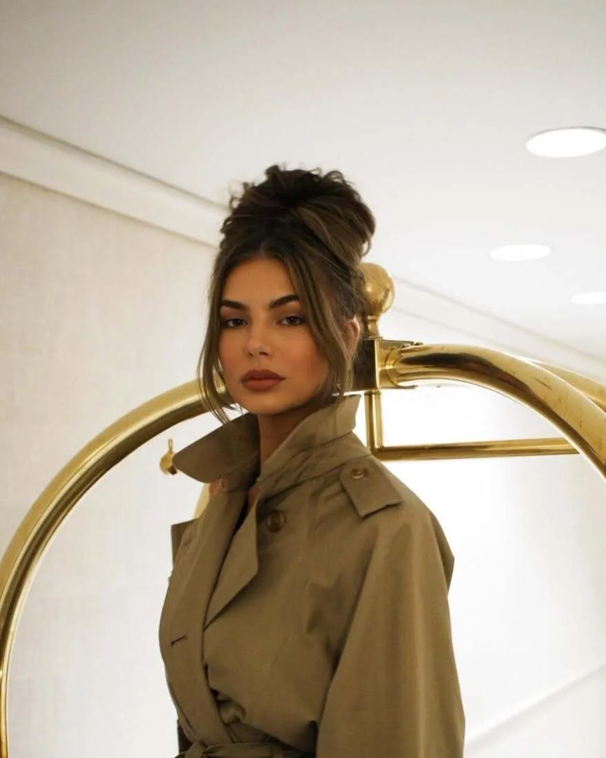 A woman with styled hair and makeup wearing a beige trench coat, standing next to a gold luggage cart in an indoor setting.