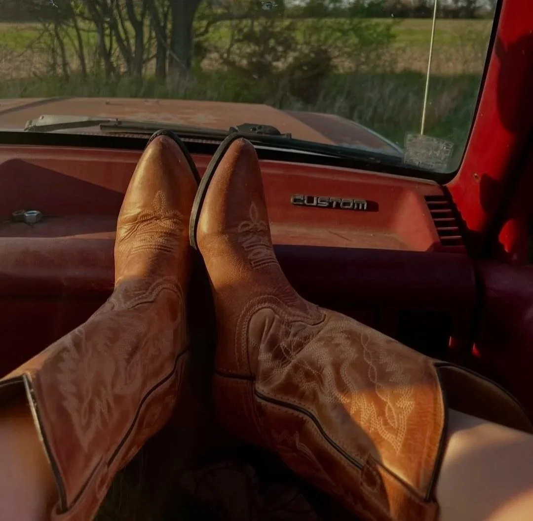 Person wearing brown cowboy boots resting their feet on the dashboard of an old red truck, with a view of trees and a field outside.
