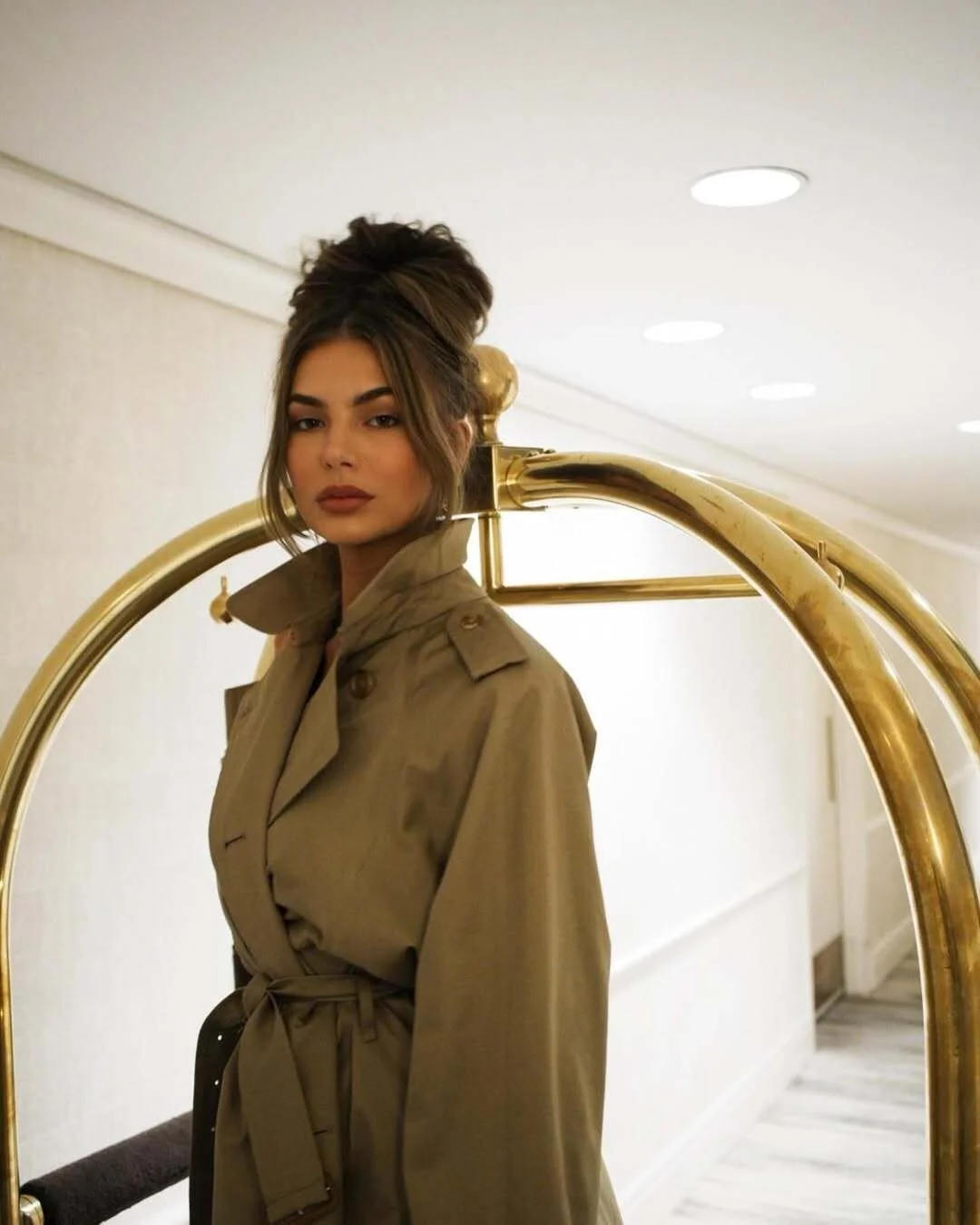Close-up portrait of woman in structured olive trench coat with neutral makeup and sleek updo in hotel hallway.