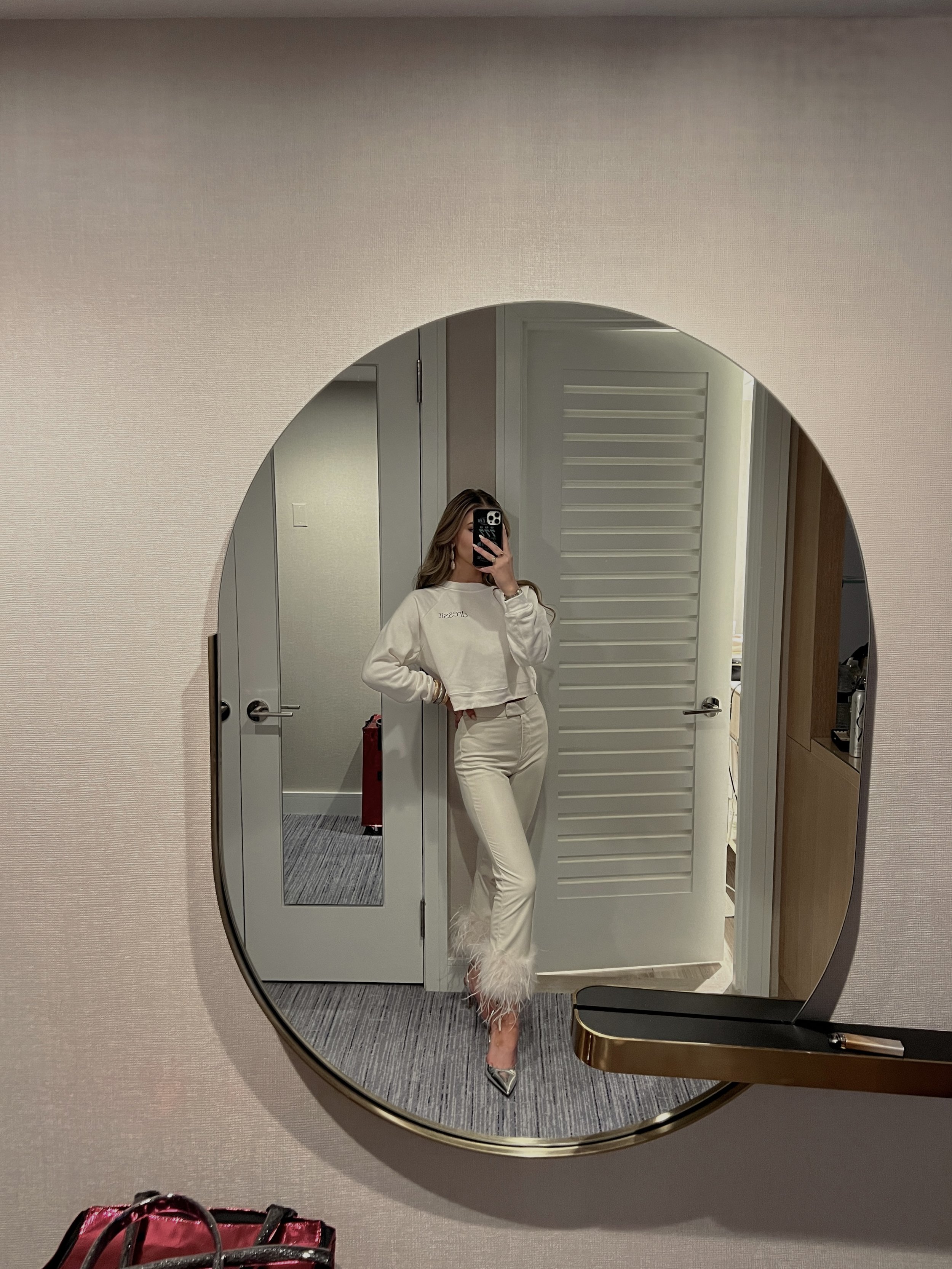 Woman taking mirror selfie in hotel room wearing cream feather-trim trousers and cropped sweater.