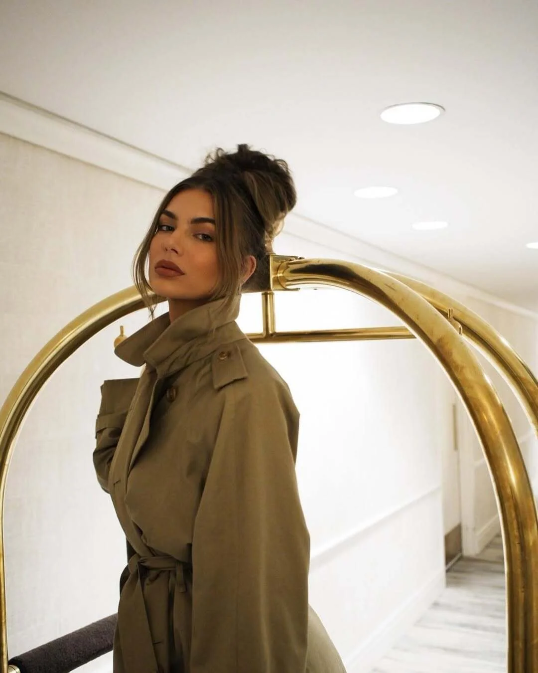 Woman in olive green trench coat with hair in an updo standing in a hotel hallway beside a gold luggage cart.