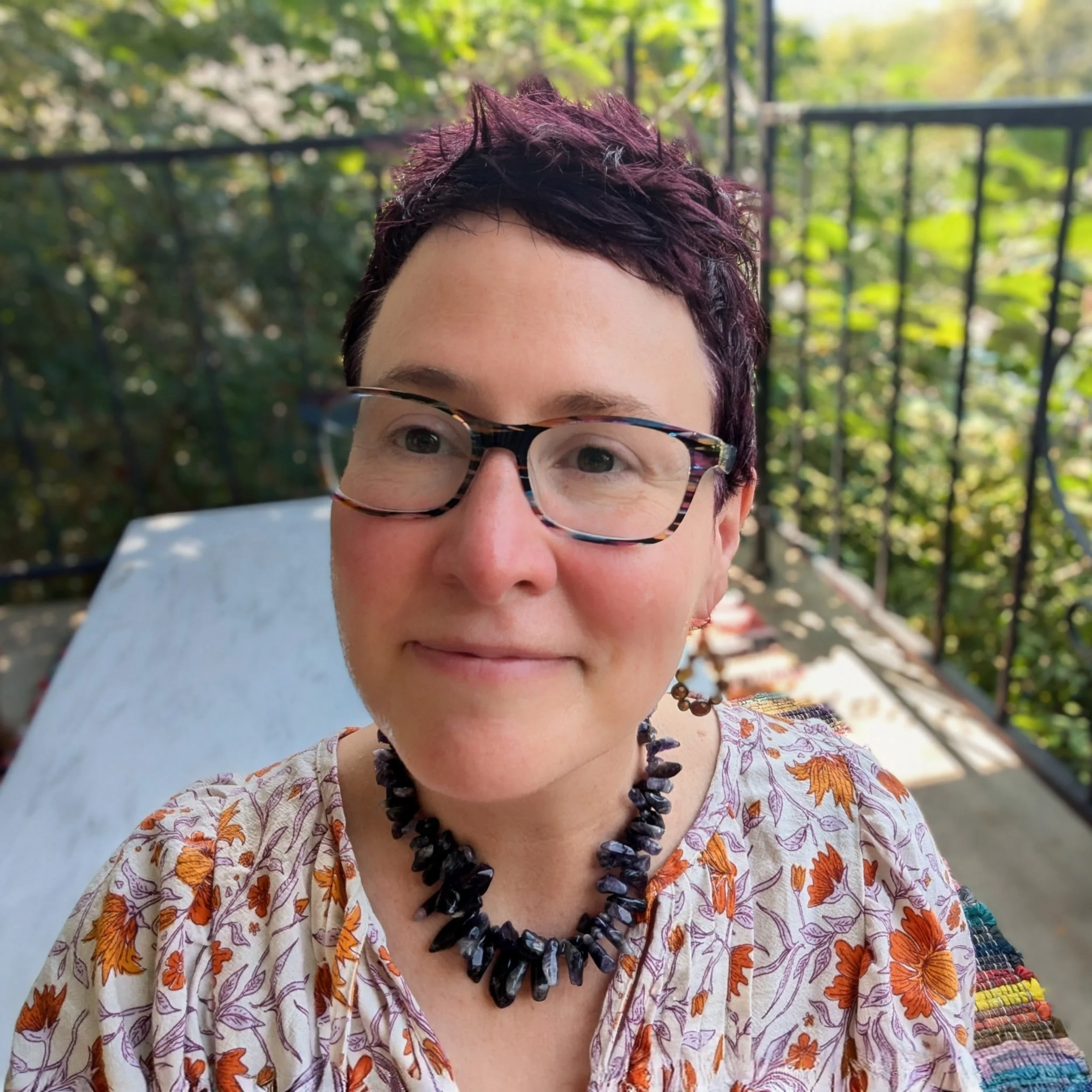 Close-up of a woman with short purple hair, glasses, a black stone necklace, and earrings, smiling outdoors on a balcony with greenery in the background.