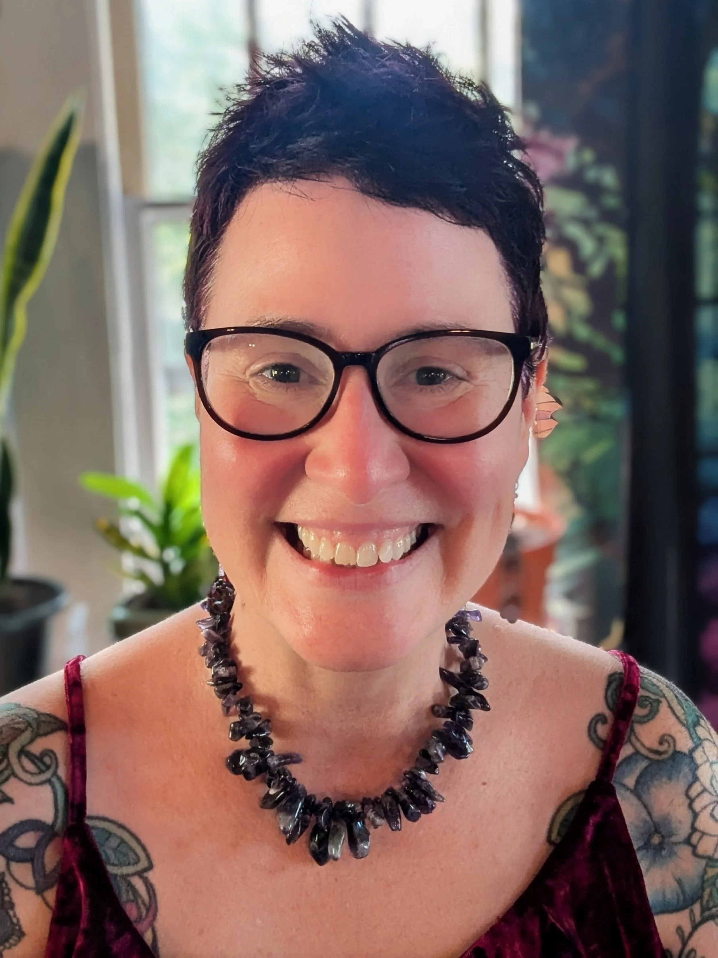 A smiling woman with short dark hair, wearing glasses, a black gemstone necklace, and tattoos on her shoulders, indoors with plants and windows in the background.