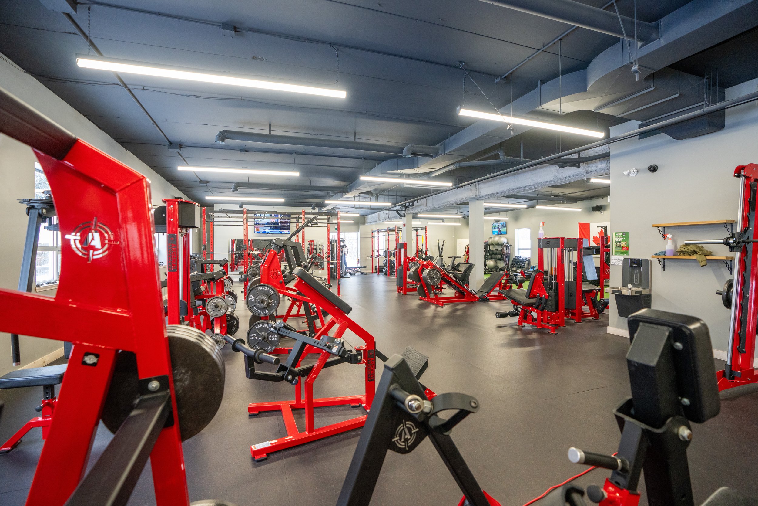 Empty gym with red fitness equipment and black flooring, large windows, overhead lighting, and a flat-screen TV on the wall.