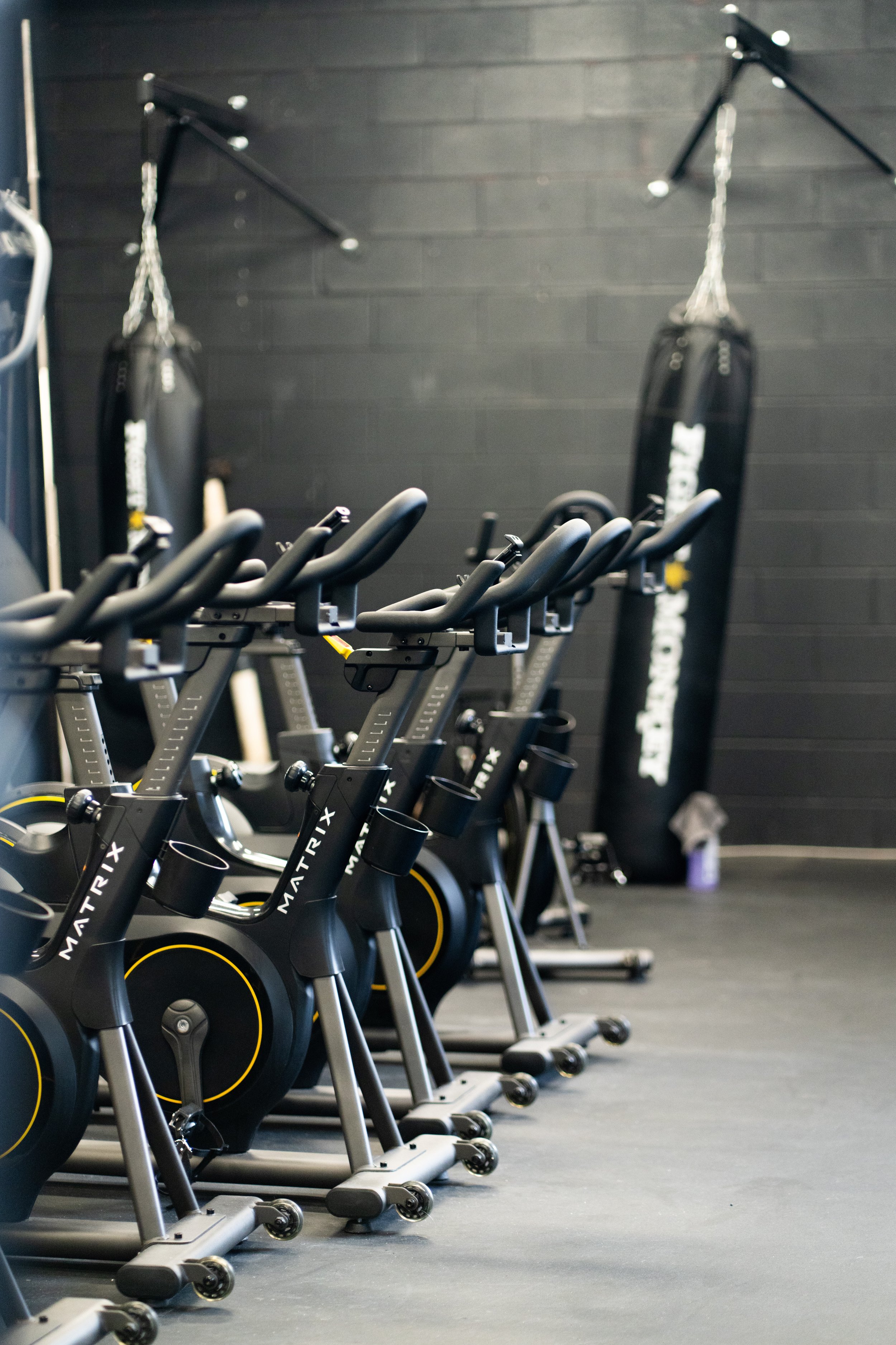 Row of black stationary exercise bikes in a gym, with punching bags hanging on the wall in the background.