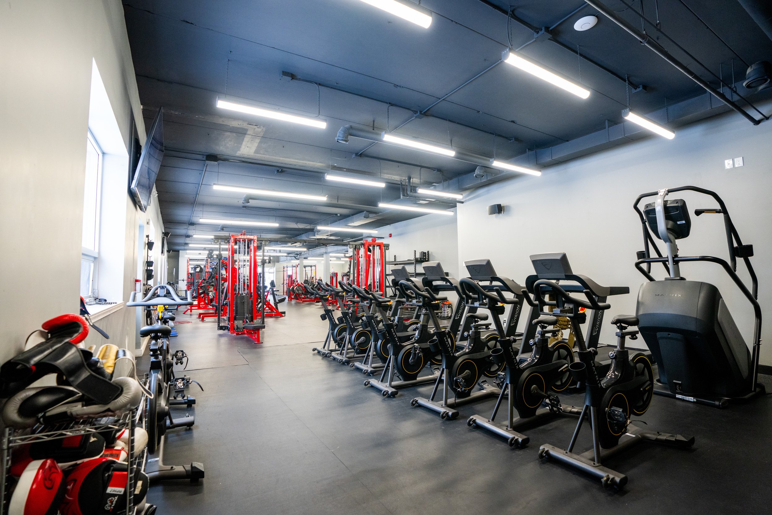 Modern gym with cardio machines like stationary bikes and an elliptical trainer, and weightlifting equipment including red and black weight racks.