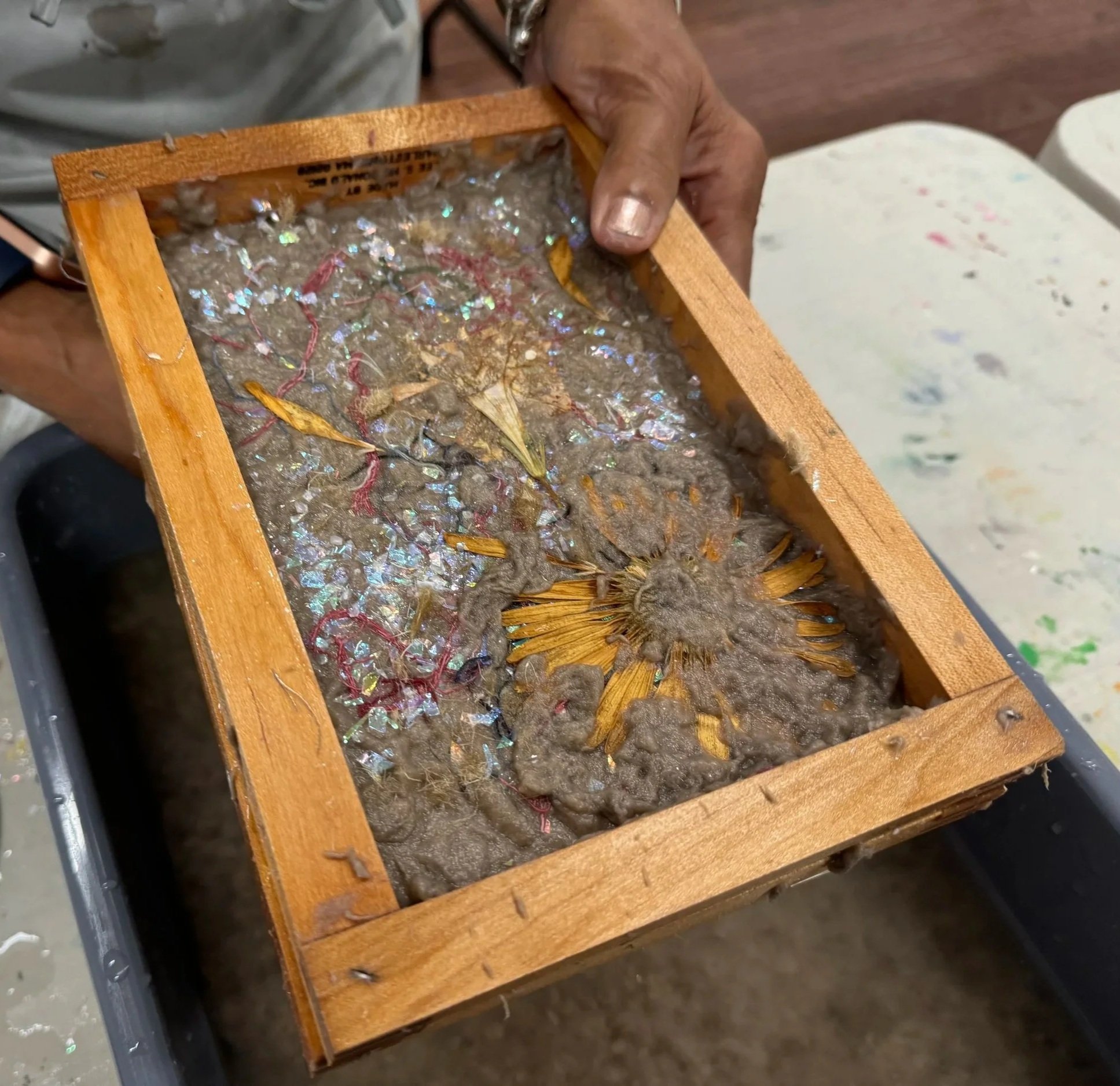 A wooden deckle filled with paper pulp, including dried flowers, leaves, and fibers.