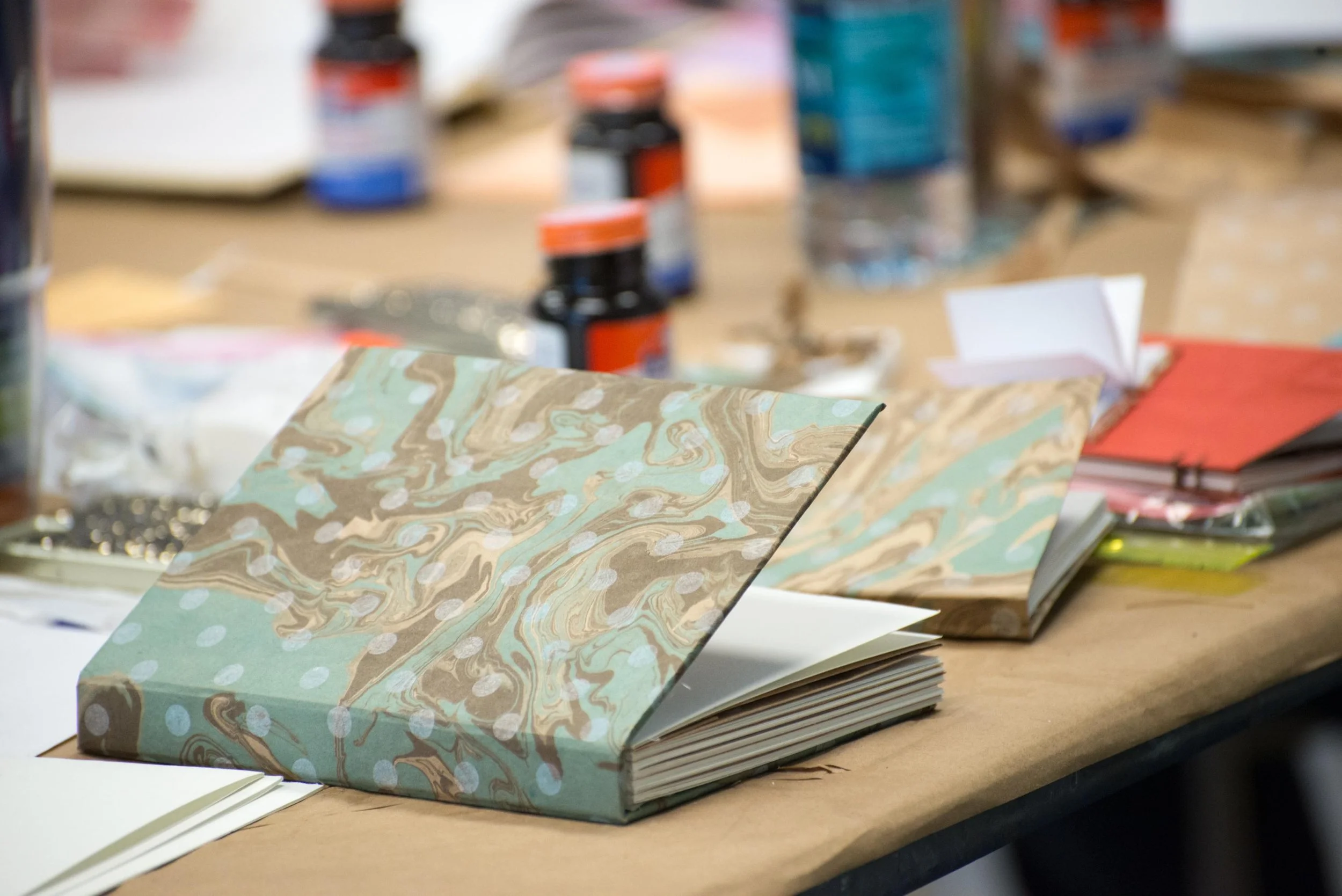 Open handmade book with colorful marble and polka dot design on the cover, resting on a cluttered wooden table with various craft supplies and papers in the background.