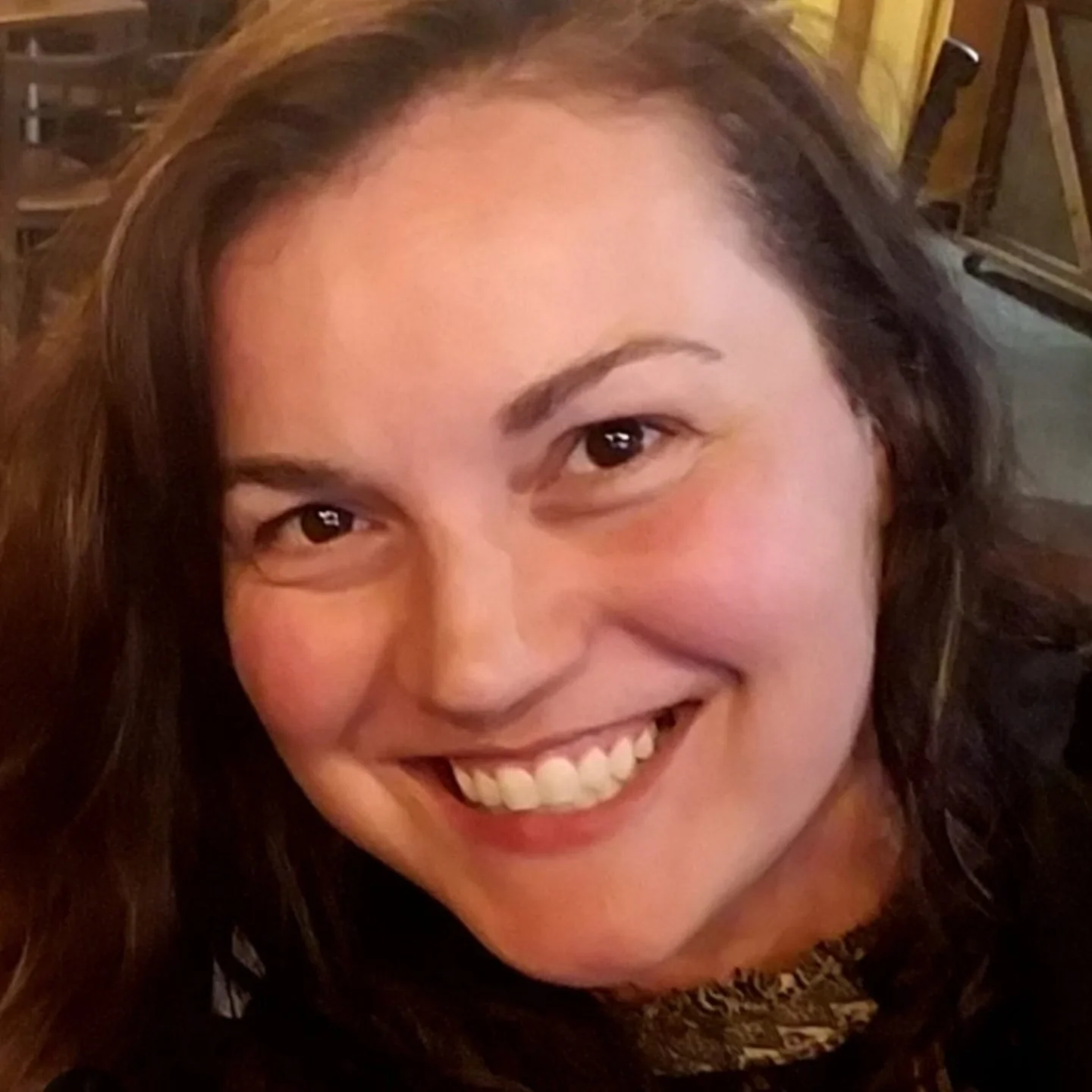 Close-up of a smiling woman with brown hair, dark eyes, and light skin in a cozy indoor setting.