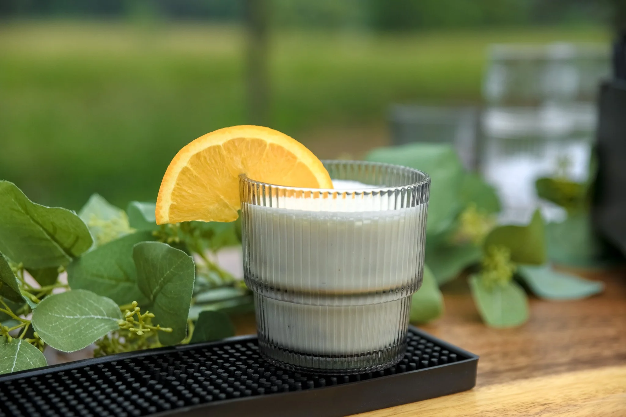 A glass of milk with a slice of orange garnish on the rim, placed on a black mat on a wooden surface, with green leaves in the background.