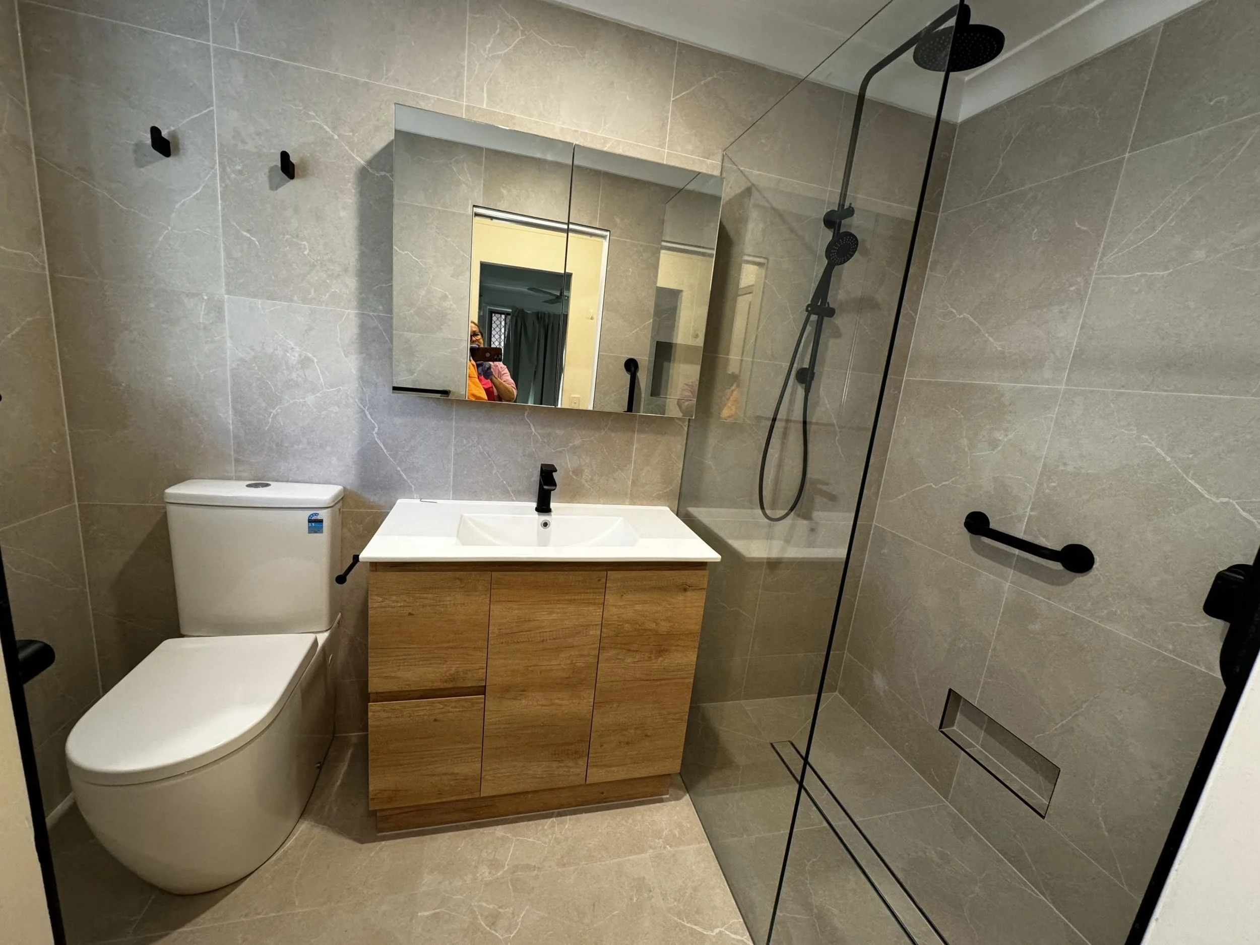 Modern bathroom with beige tiled walls and floor, a white toilet, a wooden vanity with a white sink, a rectangular mirror, and a glass-enclosed shower with black fixtures.