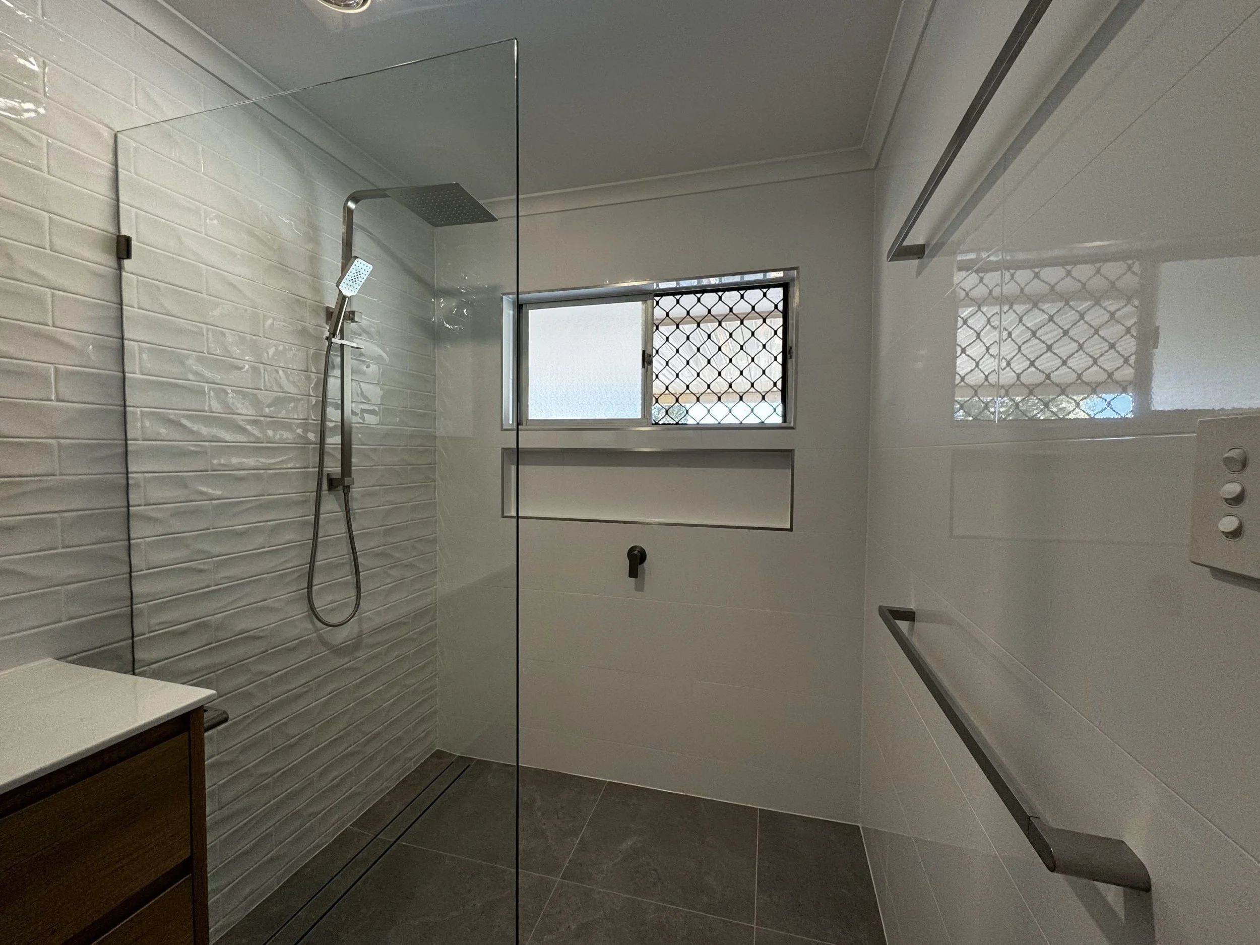 Modern walk-in shower with white textured tile wall, glass enclosure, silver shower head, small window with security bars, and dark tile flooring.